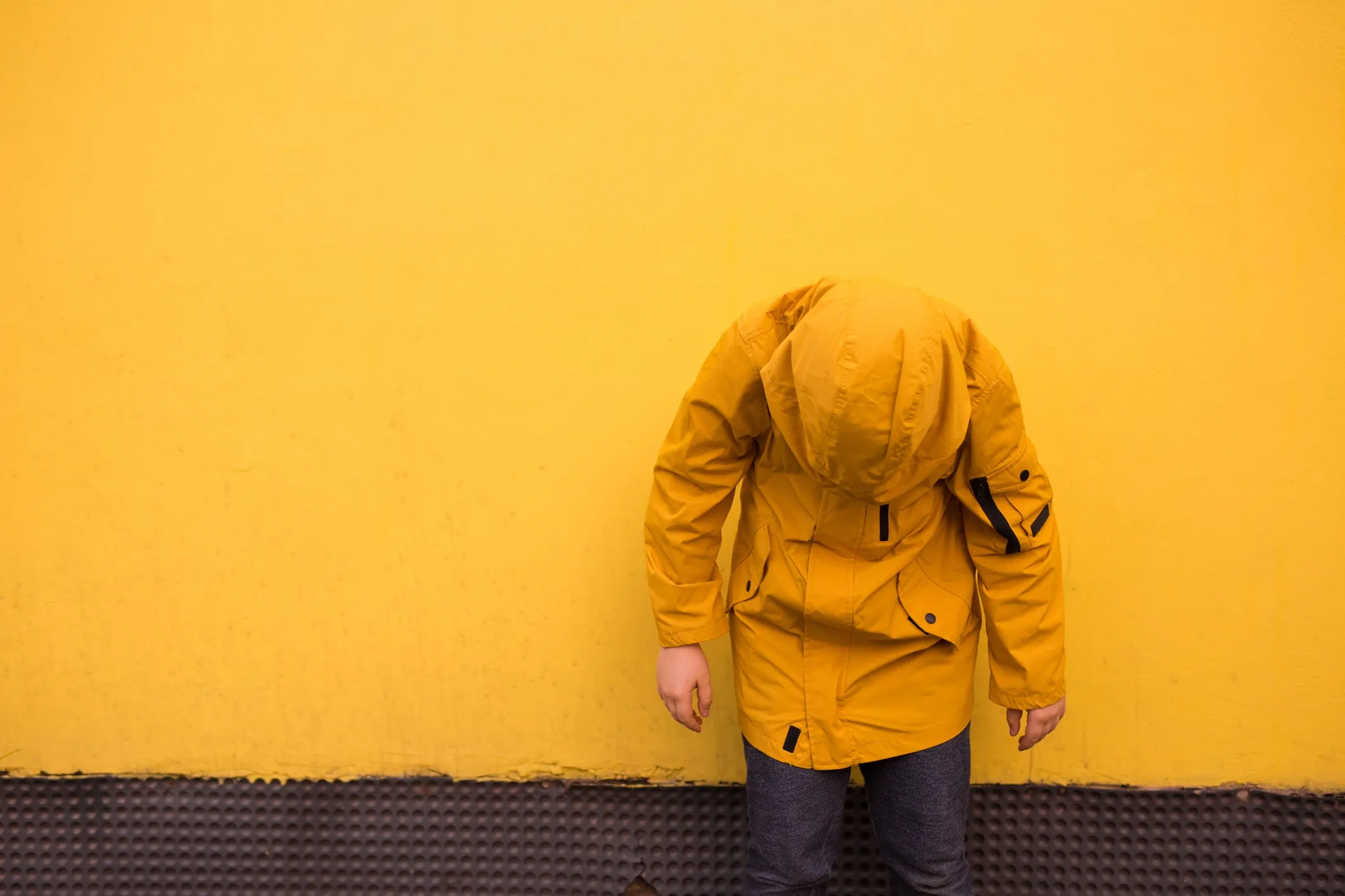 A person in a yellow jacket and dark pants standing in front of a yellow wall, with their head bowed down, obscuring their face.