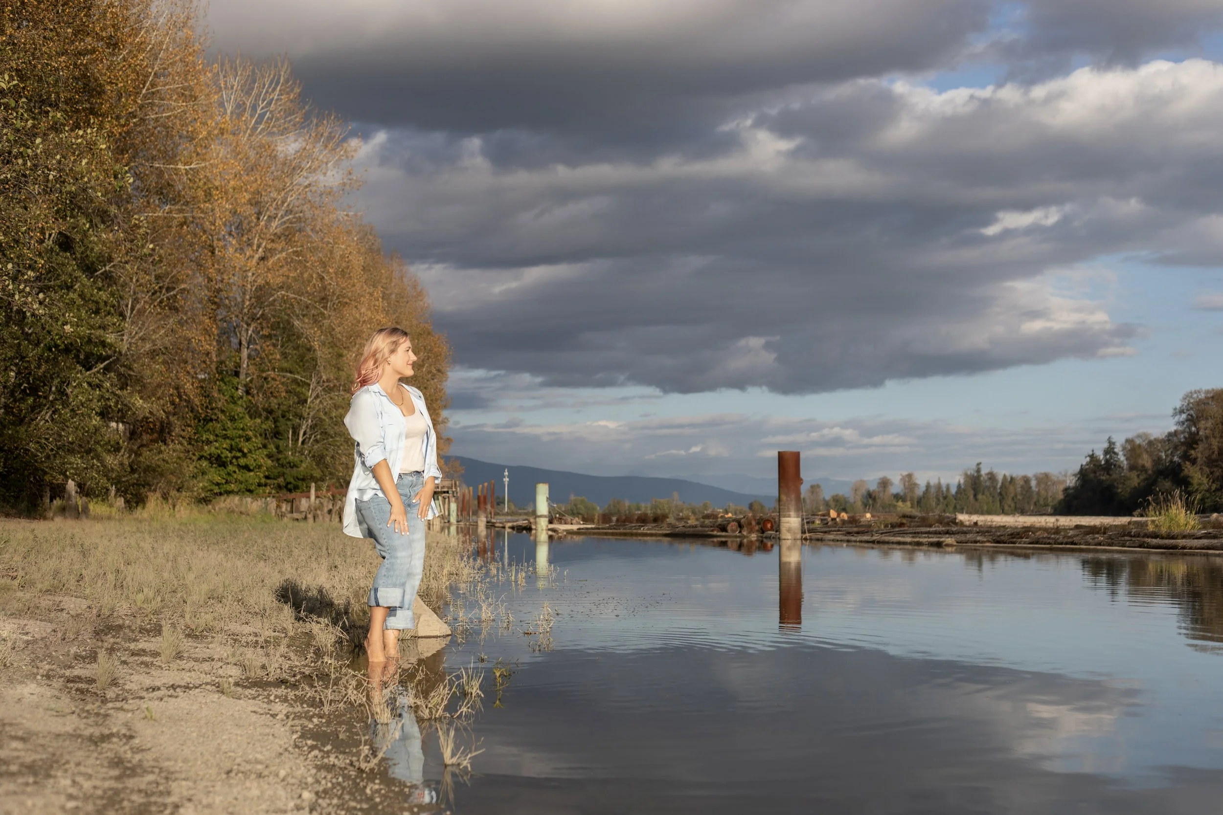 A woman in jeans and a light-colored shirt standing at the edge of a body of water, with her feet in the shallow water, looking to her right. There are trees with autumn-colored leaves on the left and a cloudy sky above. A distant mountain range is visible in the background.