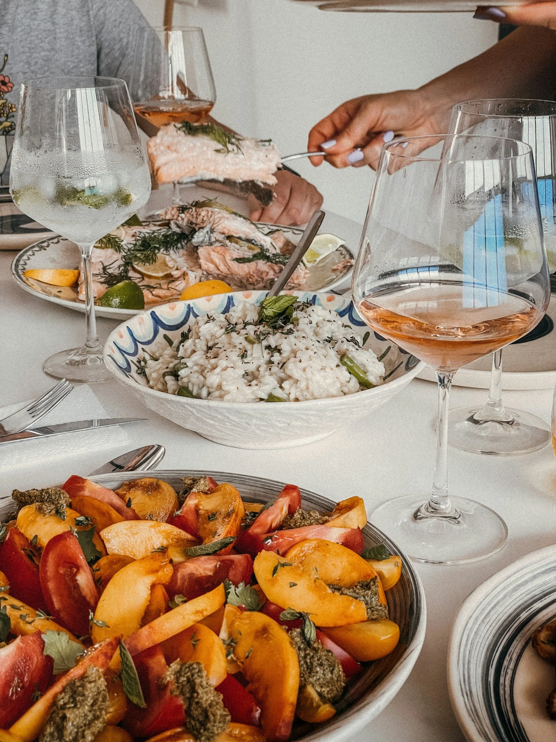 A dining table with several dishes and glasses of rosé wine, including a bowl of roasted or grilled vegetables, a plate of fresh fish garnished with herbs, and a salad with peaches and herbs.