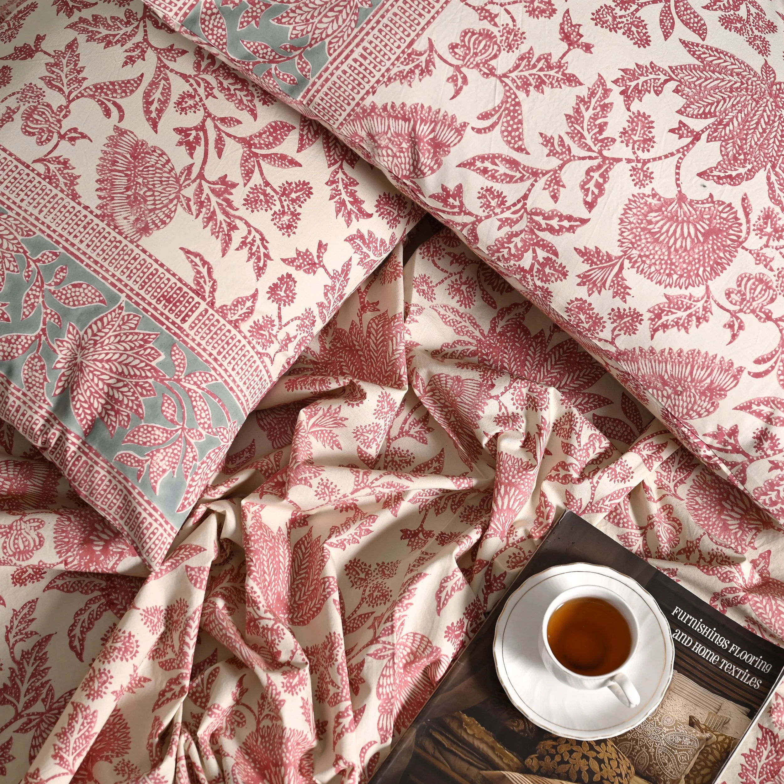 Pink and white floral patterned bed linens and pillows with a cup of tea on a saucer and a magazine.