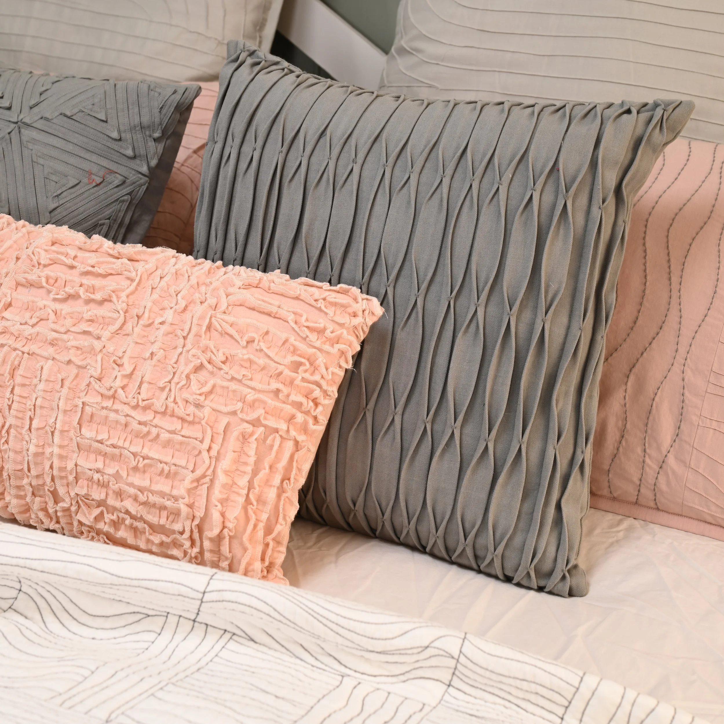 Close-up of decorative pillows on a bed, including a pink frilly pillow, a gray pillow with pleated design, and a light pink pillow with embroidery.