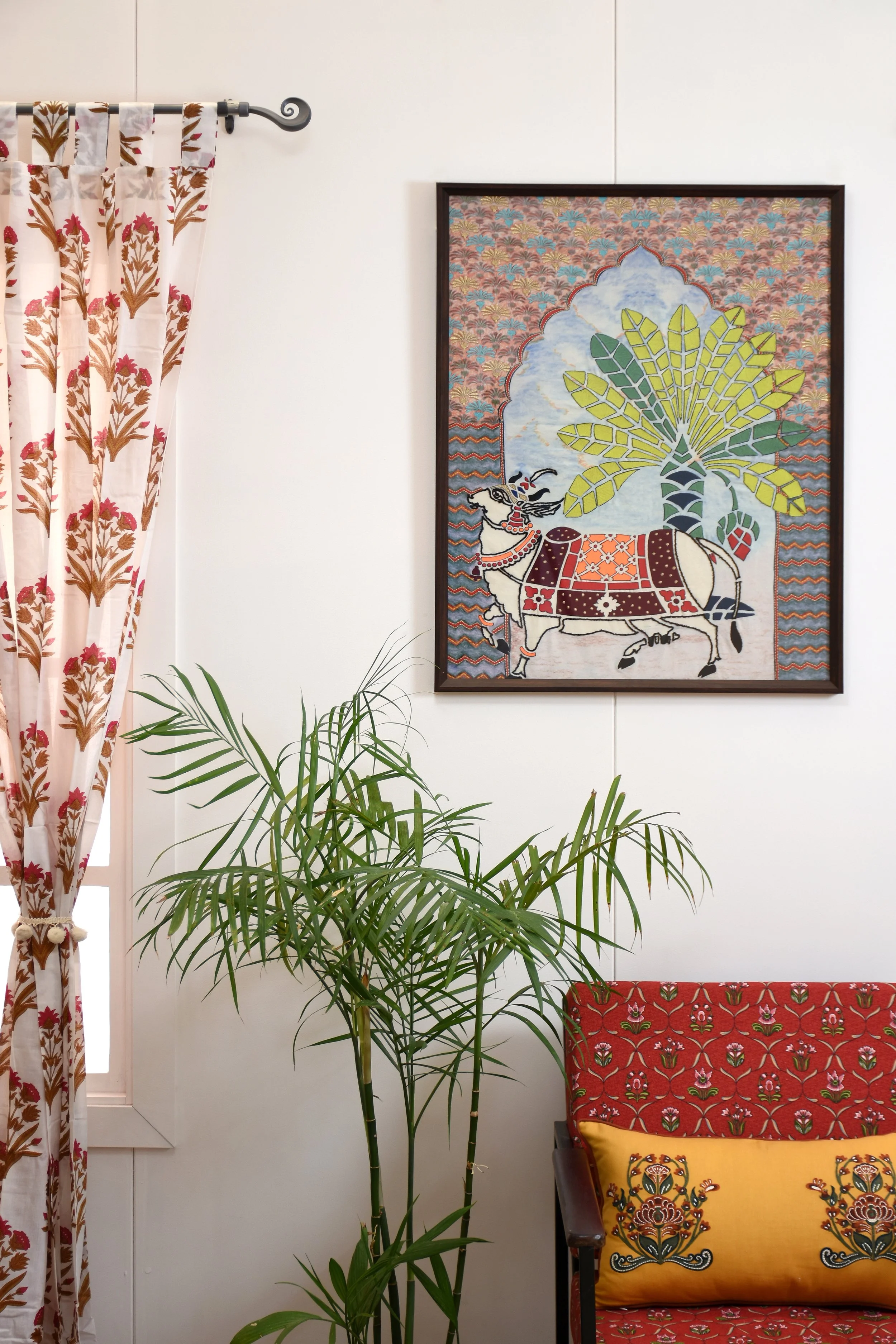 Interior scene with a colorful tapestry on the wall depicting a cow standing under a tree, a window with floral curtains on the left, a green potted plant in the foreground, and a red and yellow patterned chair with floral pillows on the right.