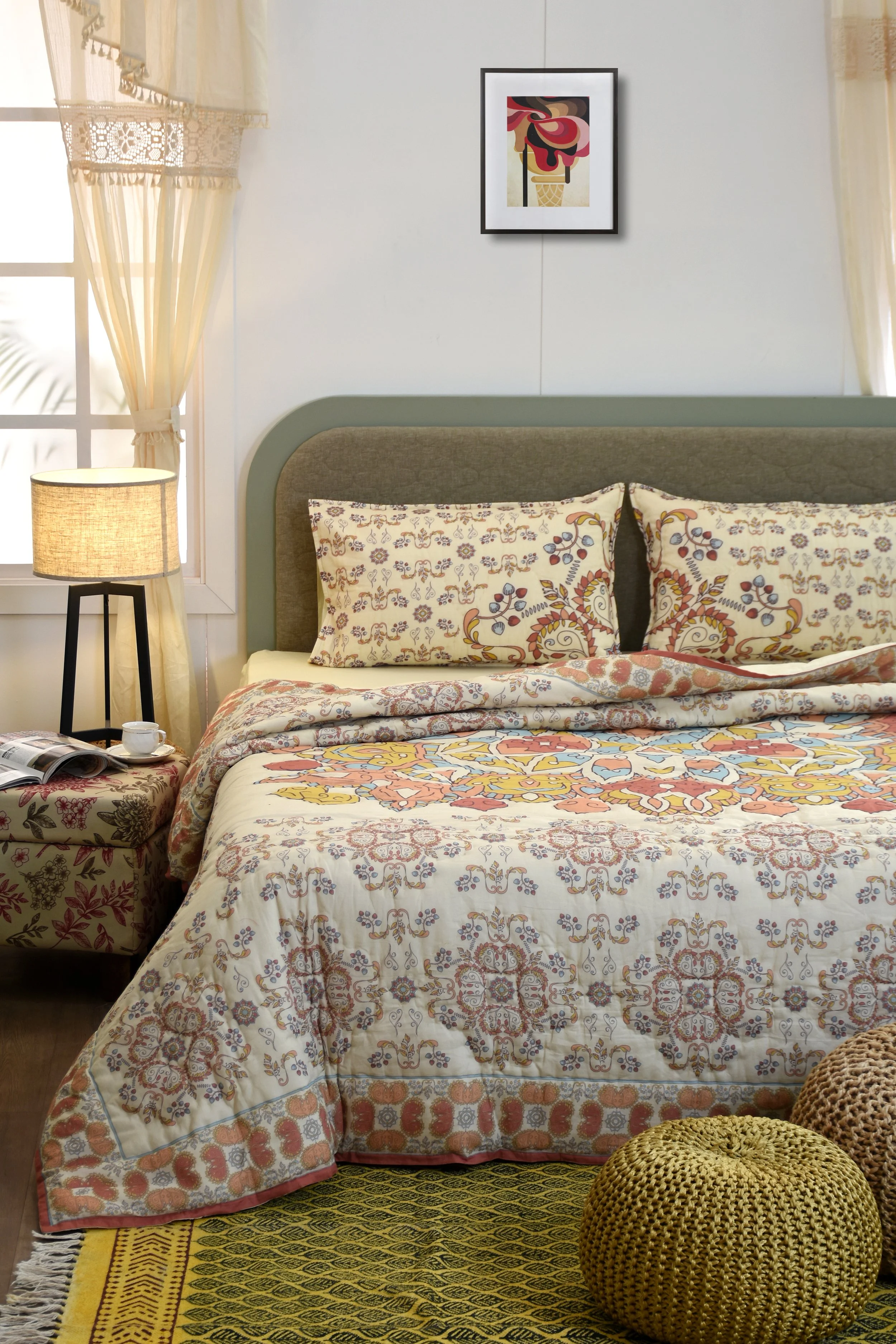 A neatly made bed with ornamental pillows and a patterned bedspread in a sunlit bedroom. There is a bedside table with a lamp, a cup, and an open magazine. A framed abstract artwork hangs above the bed, and yellow curtains with lace trim are on the w