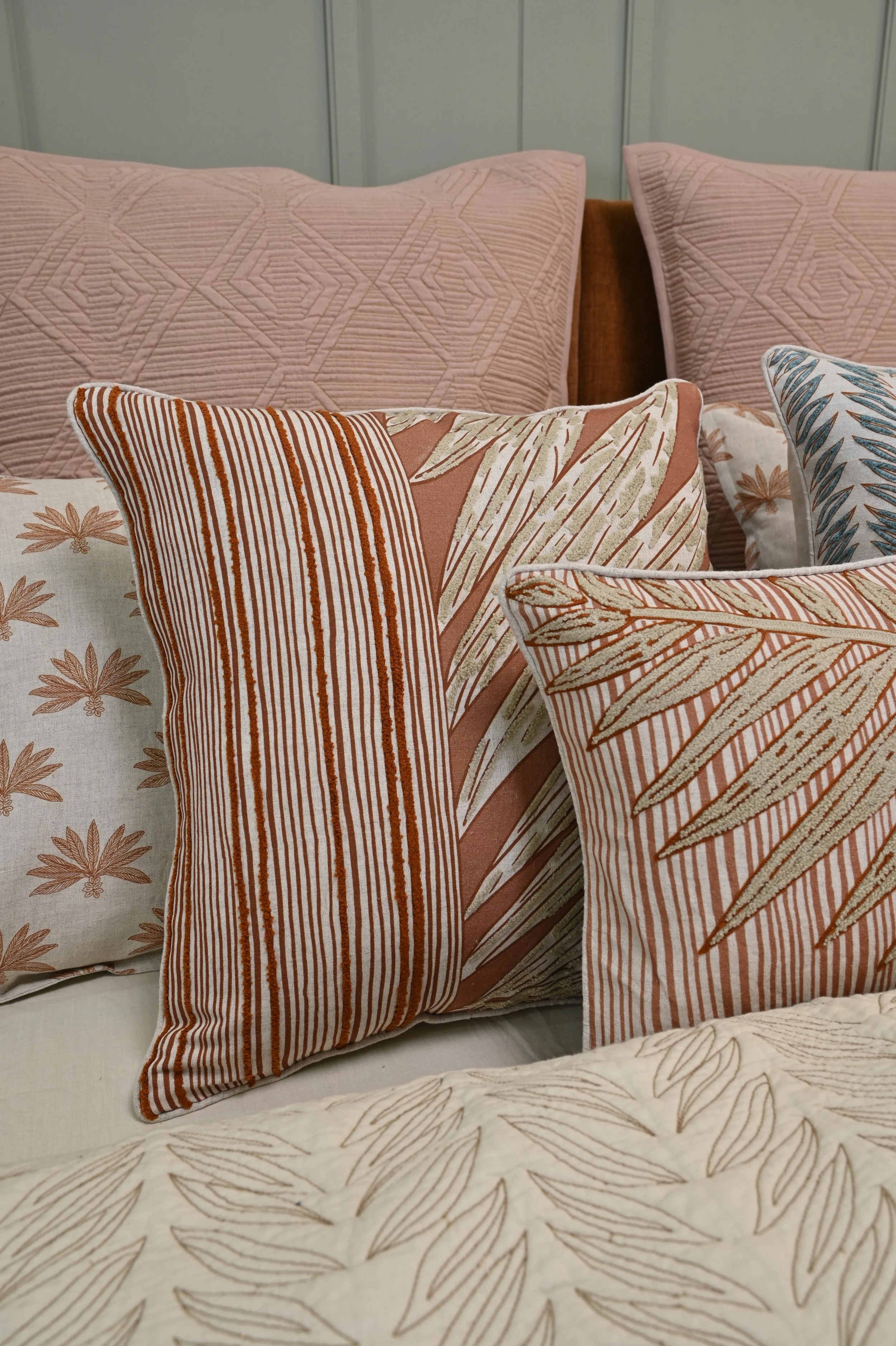 Close-up of decorative pillows with geometric and botanical patterns on a bed, featuring colors like pink, beige, peach, and blue.