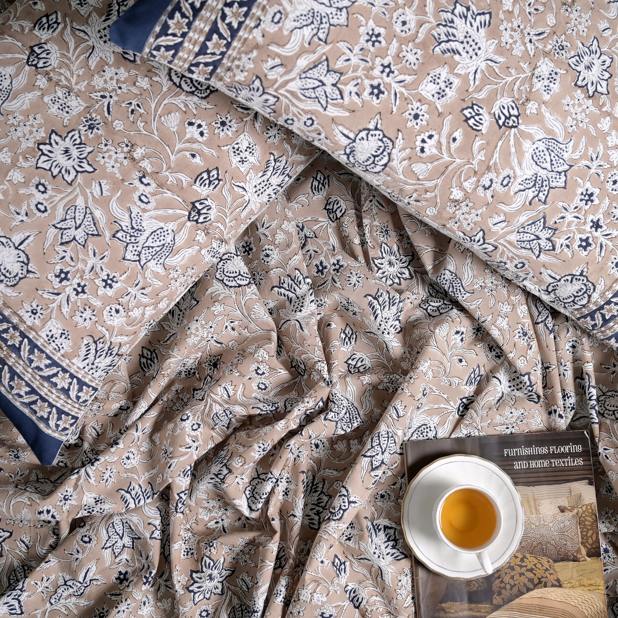 Display of beige fabric with a navy and white floral pattern, draped and folded, with a tea cup and magazine on top. The magazine's title reads 'Furnishings Flooring and Home Textiles'.