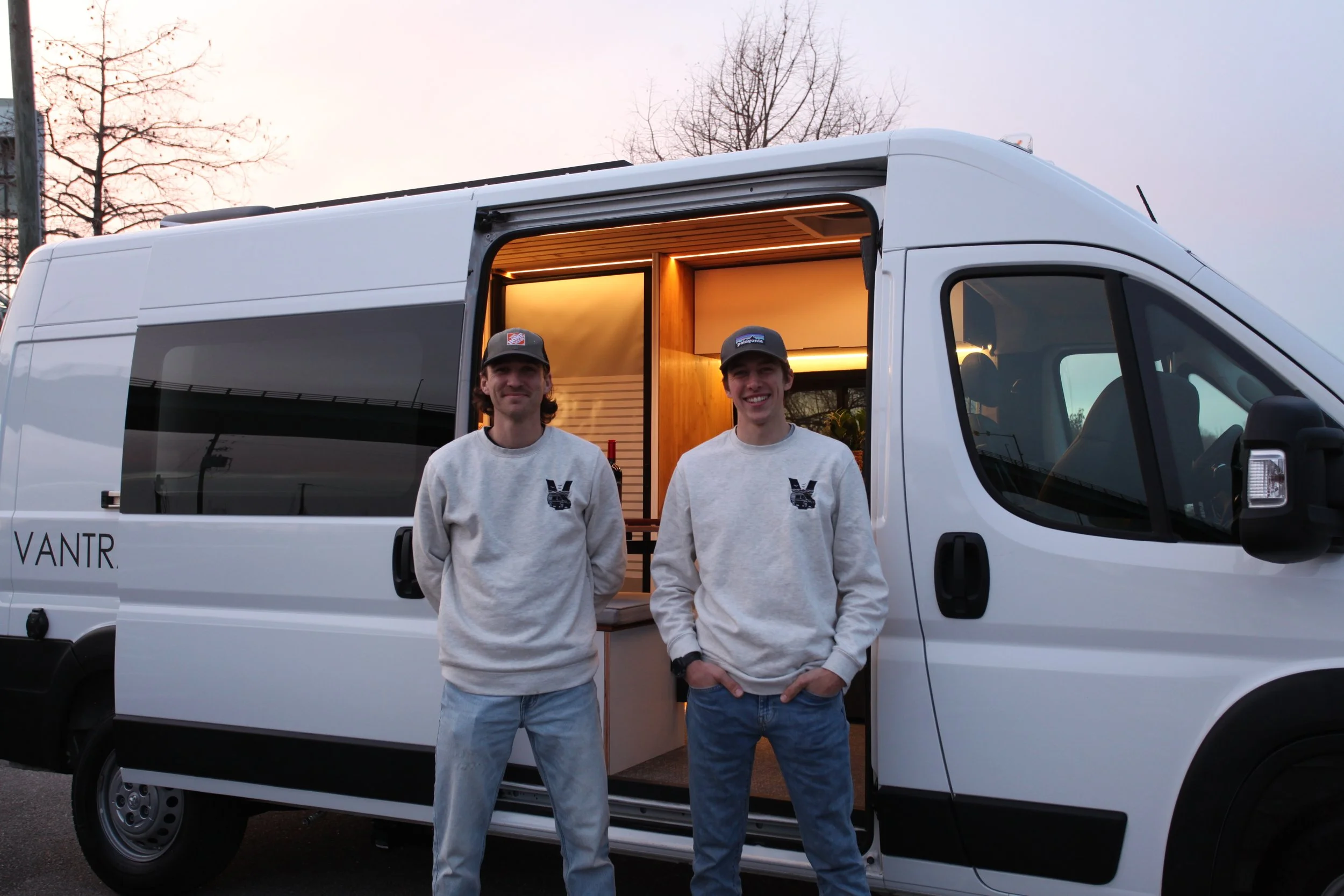 Two young men in gray sweatshirts and baseball caps standing in front of a white van with its side door open, revealing a wood-paneled interior, at sunset.