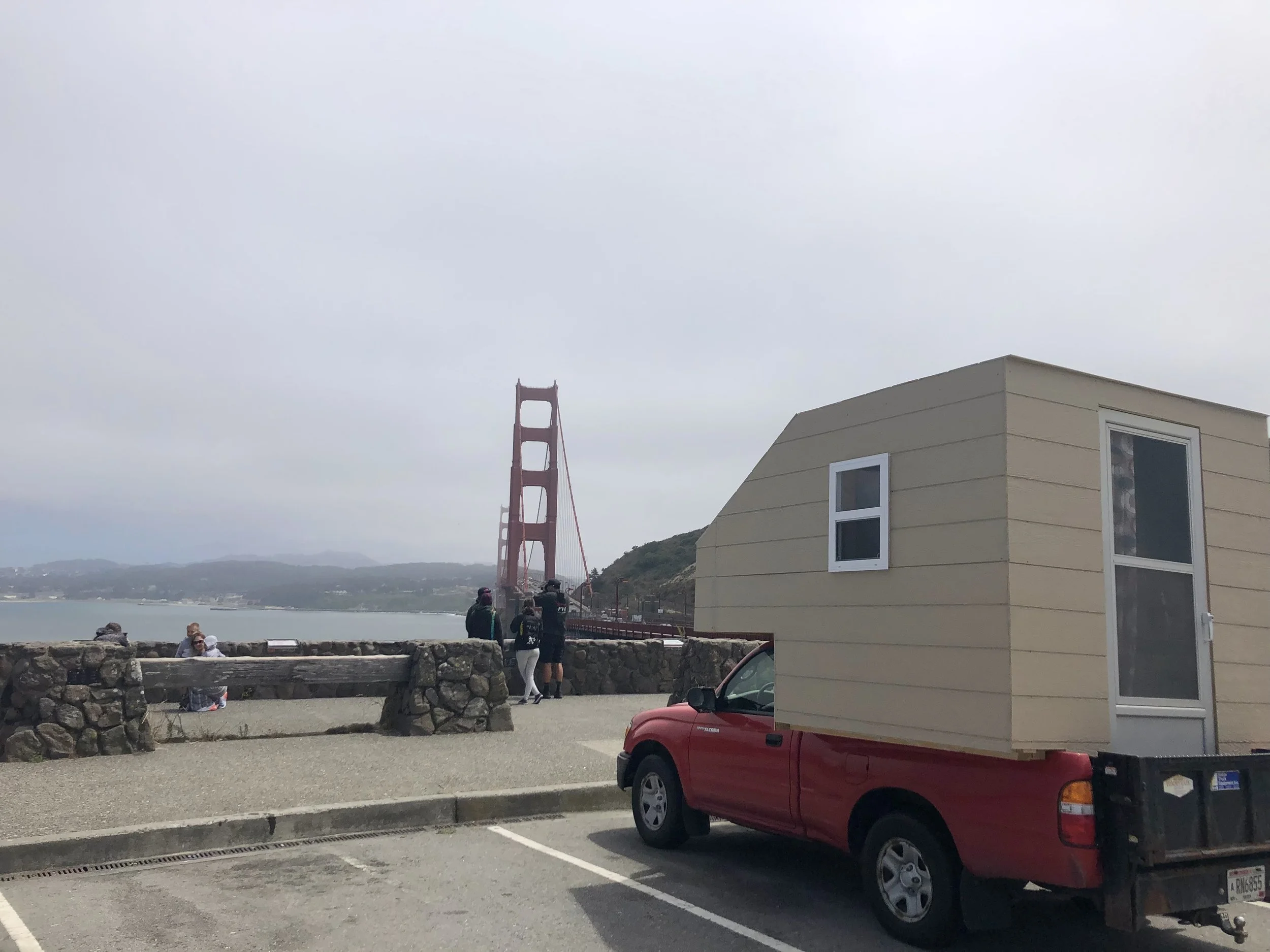 A red car parked in a parking lot with a beige tiny house on its back. In the background, the Golden Gate Bridge is partially visible through the cloudy sky, with several people standing near the water and a stone barrier along the shoreline.