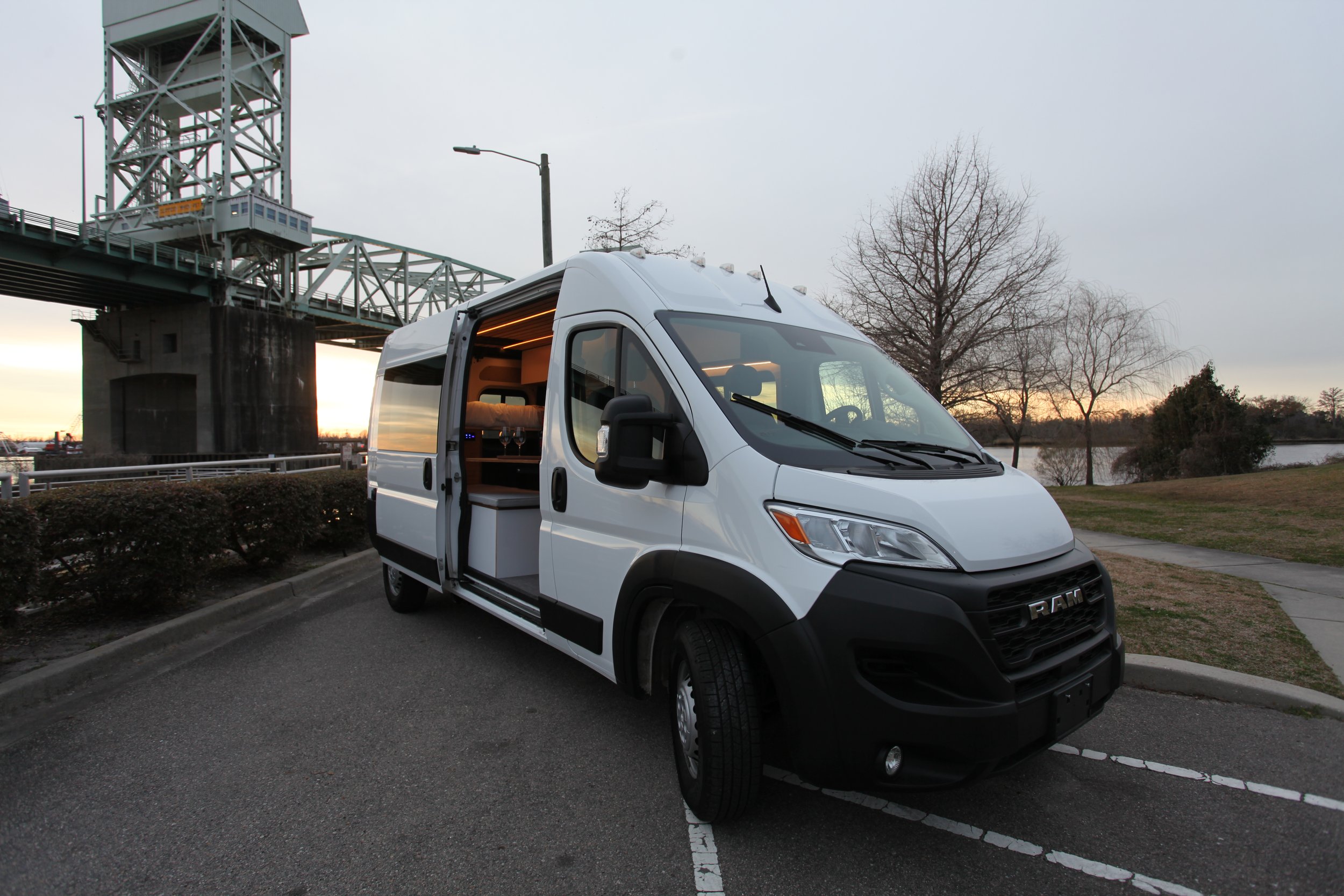 White delivery van with open sliding door parked on a street near a park, with leafless trees, a river, and a bridge at sunset in the background.