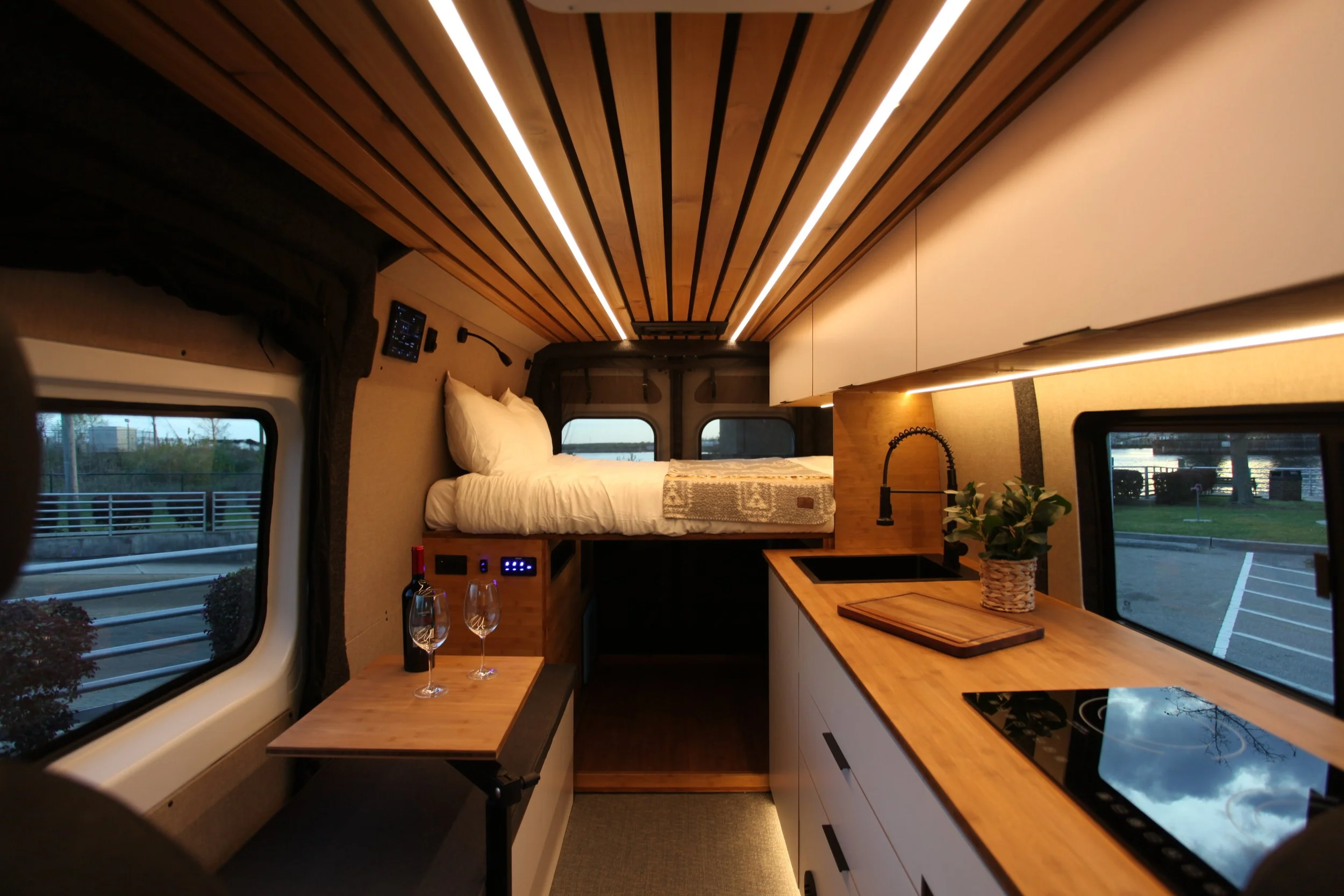 Vantra camper van interior with bed platform, kitchen galley, wood ceiling slats, custom cabinetry, and large rear windows.