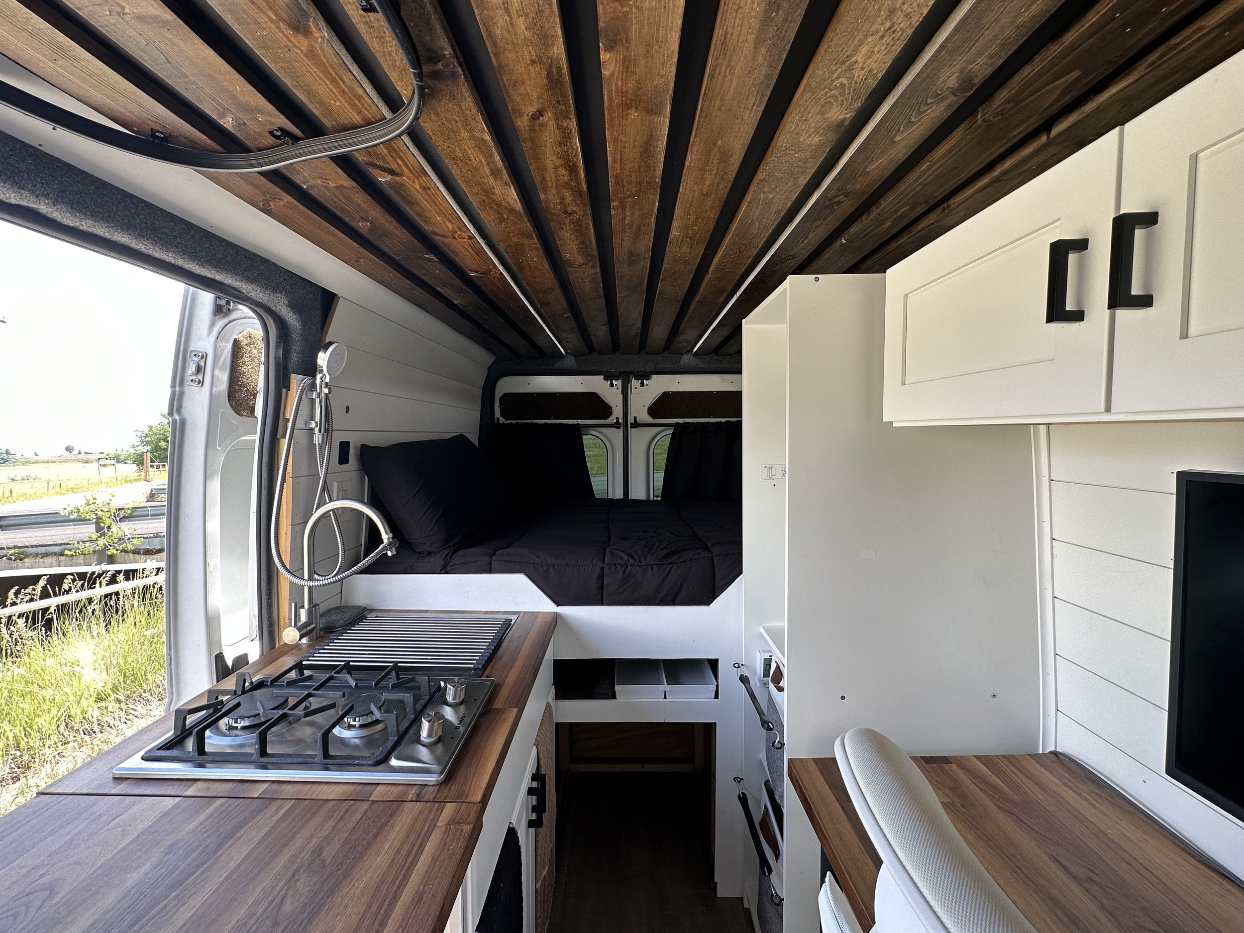 Interior of a tiny camper van with a small kitchen area, black stove with burners, wooden countertop, white cabinetry, a bed with black bedding, and ceiling made of wooden planks.