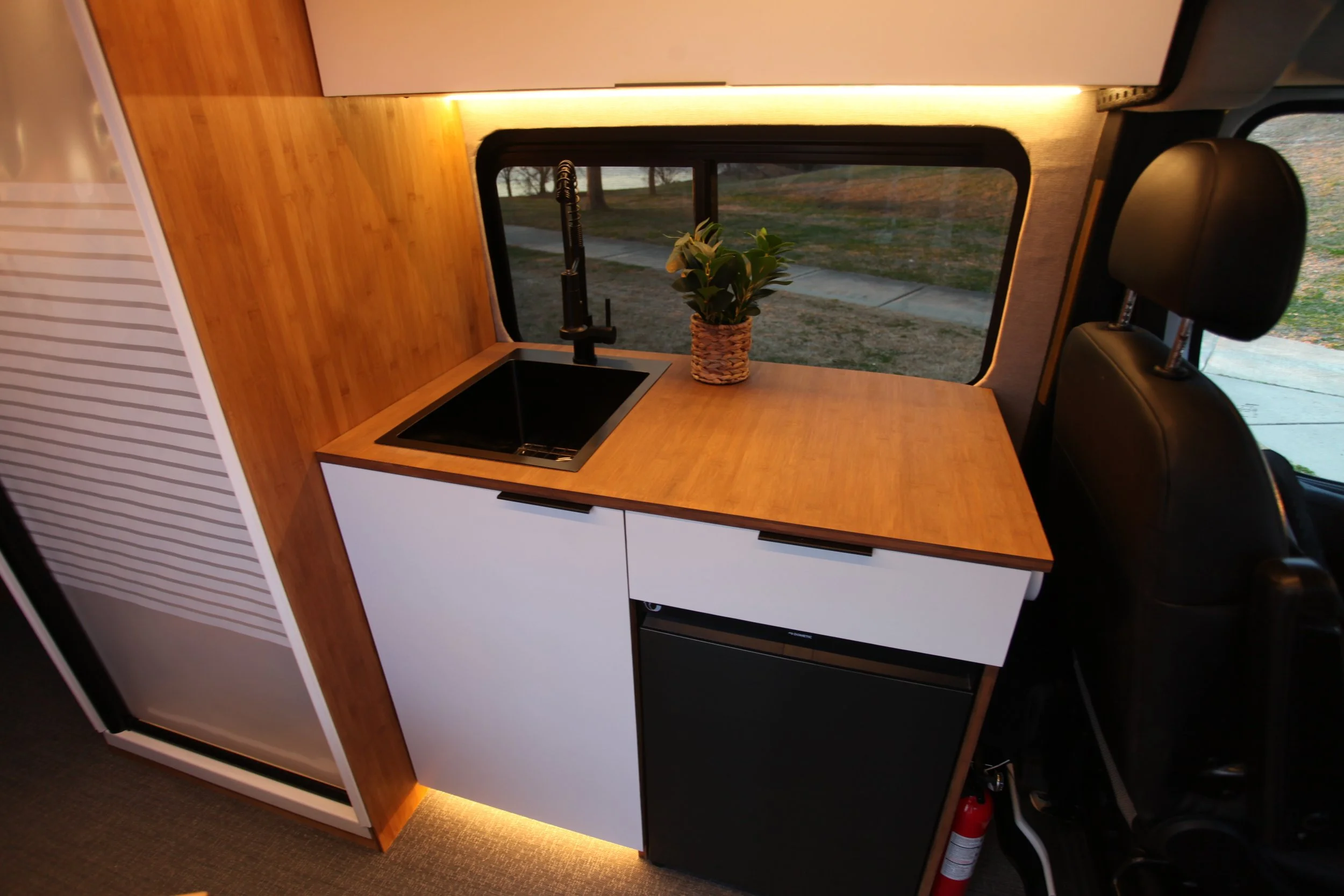 Small kitchen area inside a van or camper, with a black sink, wooden countertop, a potted plant, and a window with a view of the outdoors.
