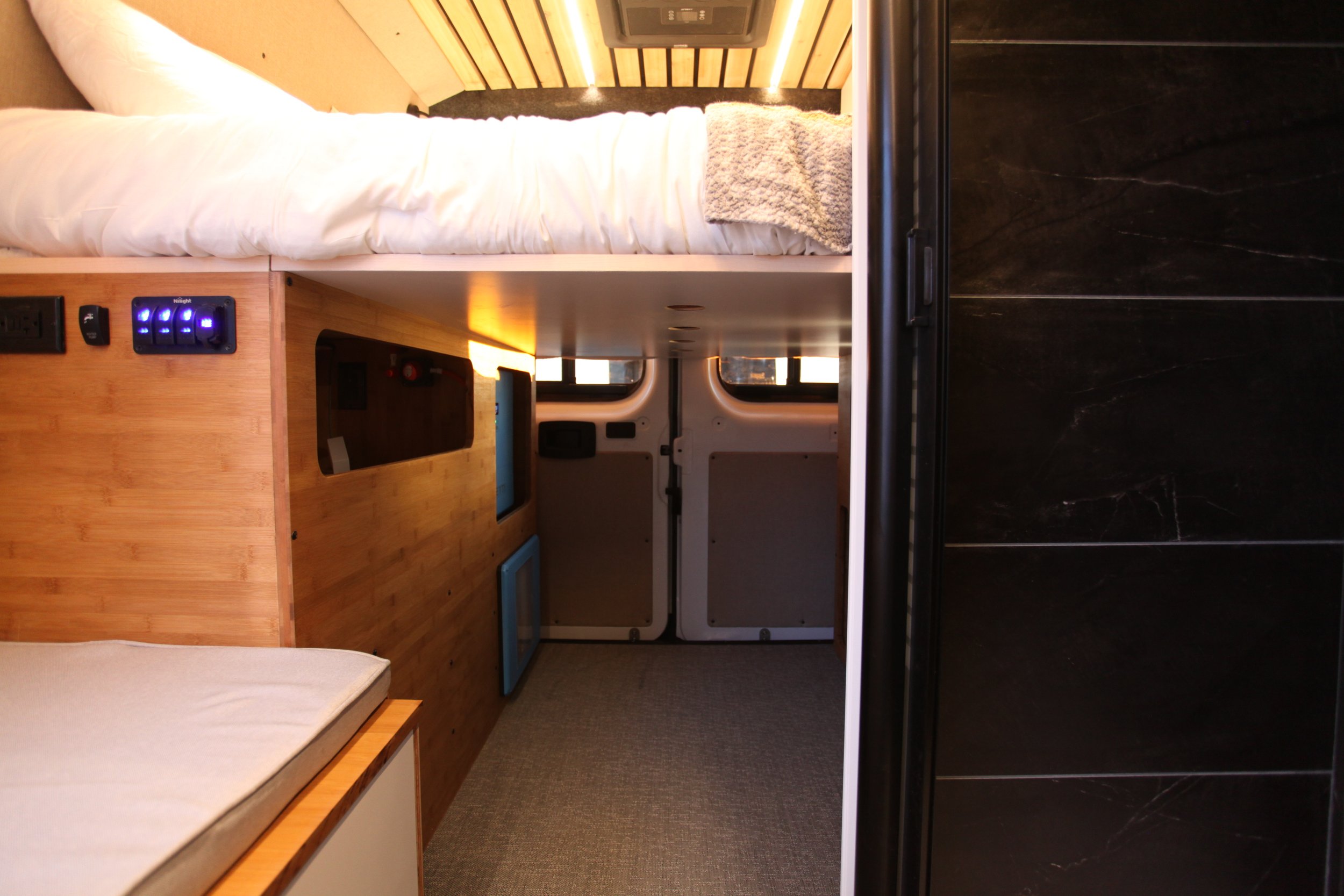 Interior of a compact living space, showing a bed with white bedding and a towel, wooden accents, a small refrigerator, and black wall tiles, with a door leading outside.