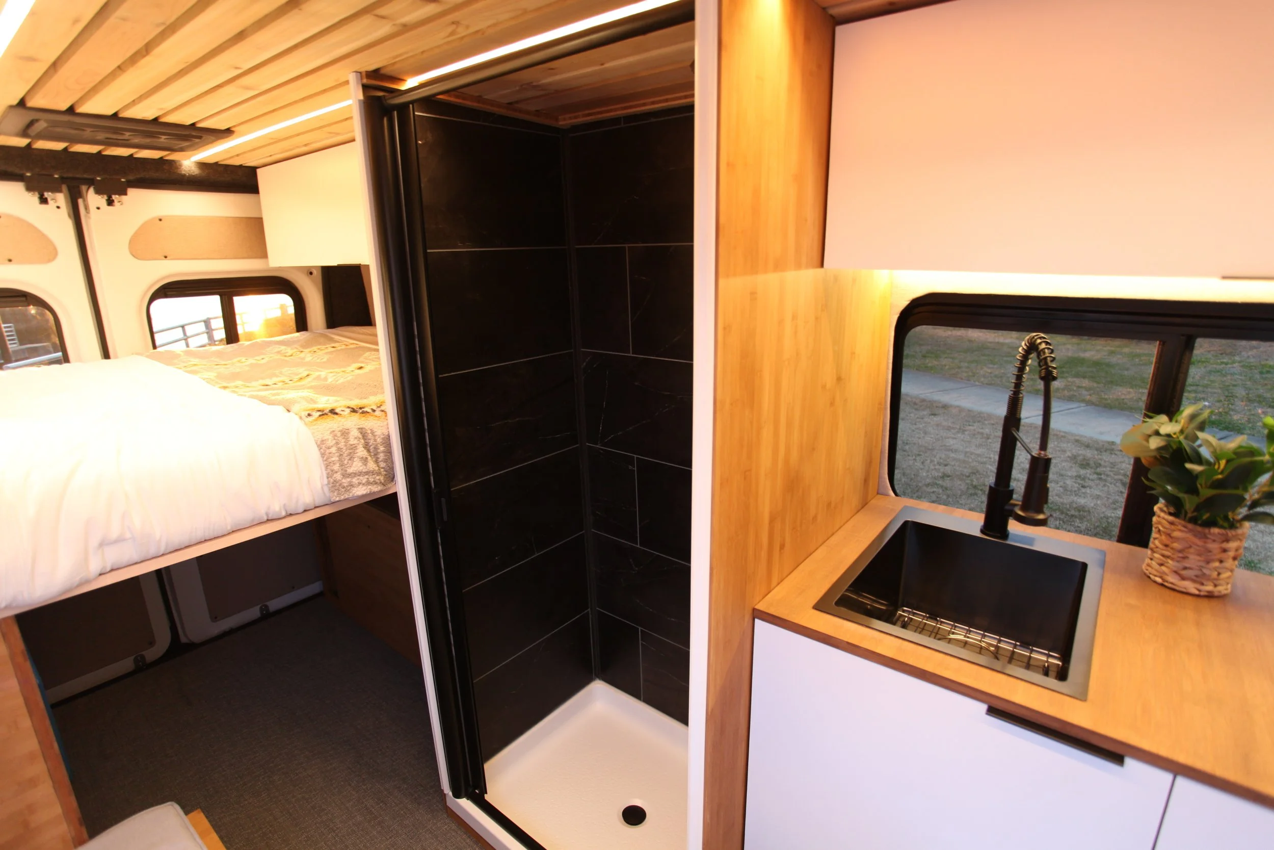 Interior of a small RV or camper with a sleeping area, compact black shower stall, and a small kitchen sink with a plant on the countertop.