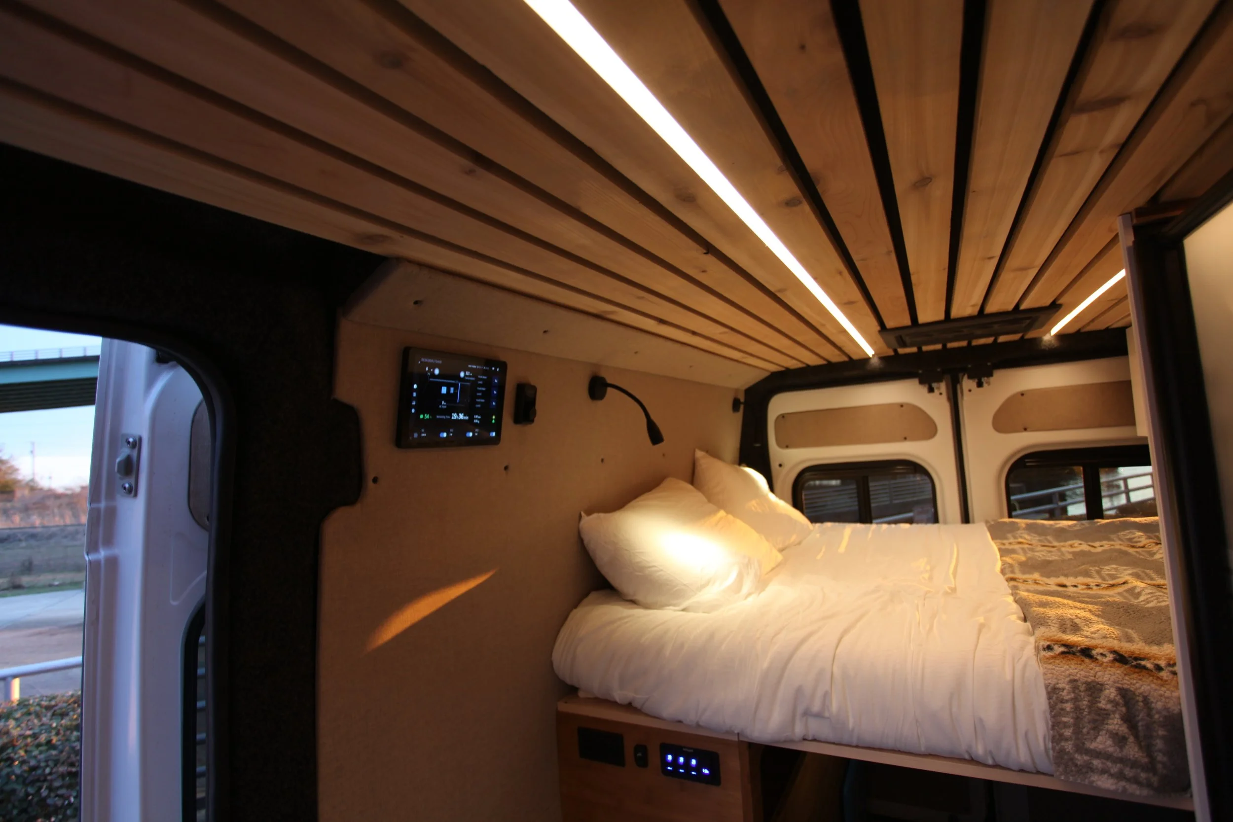 Interior of a camper van with a bed, wooden ceiling, and electronic control panel