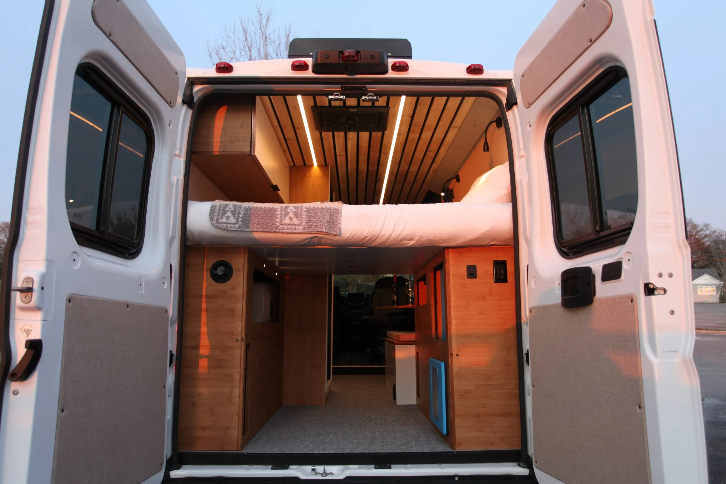 View of the interior of a camper van with a bed on the upper level and wooden cabinetry below, seen through open rear doors.