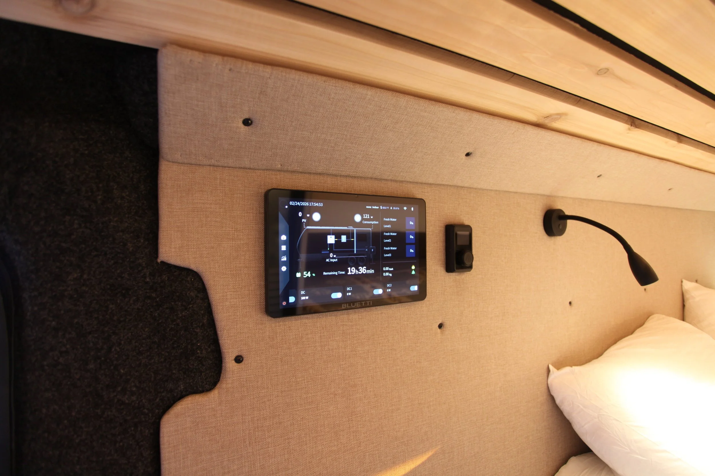 Close-up of a beige upholstered headboard with a built-in digital control panel, a black reading light, and a white pillow, in a hotel room.