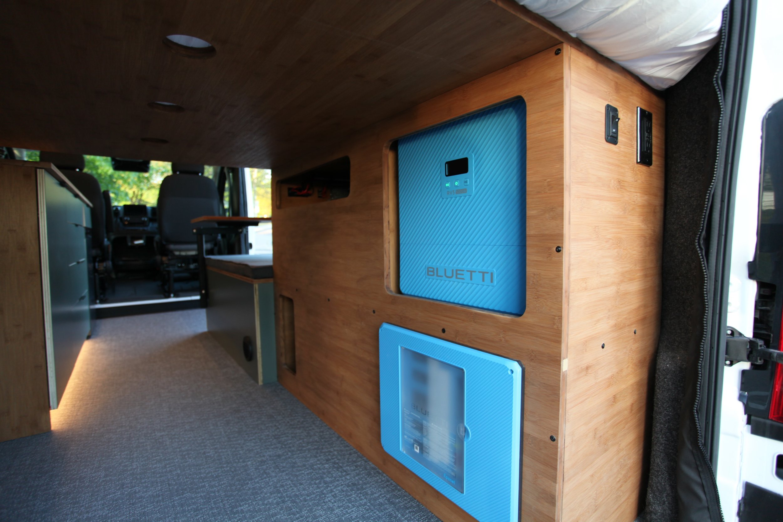 Interior of a camper van with wooden cabinetry, blue Bluetti portable power station, and a seating area with a window showing greenery outside.
