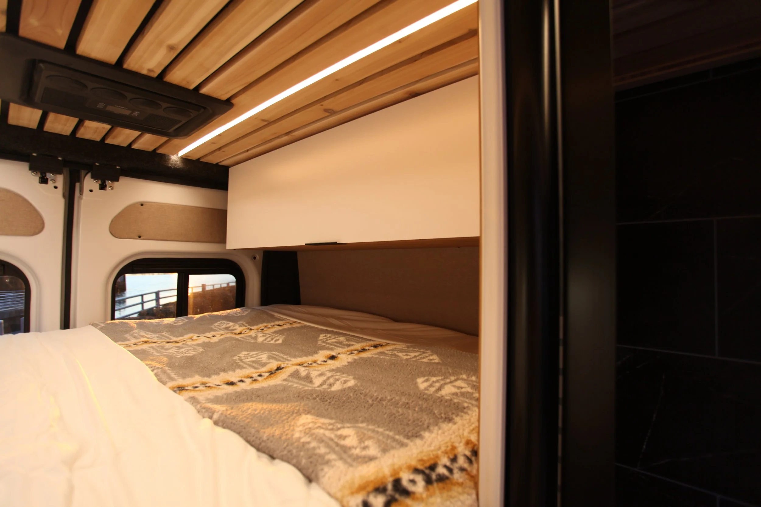 Interior of a camper van with a bed, windows, and wooden ceiling panels.