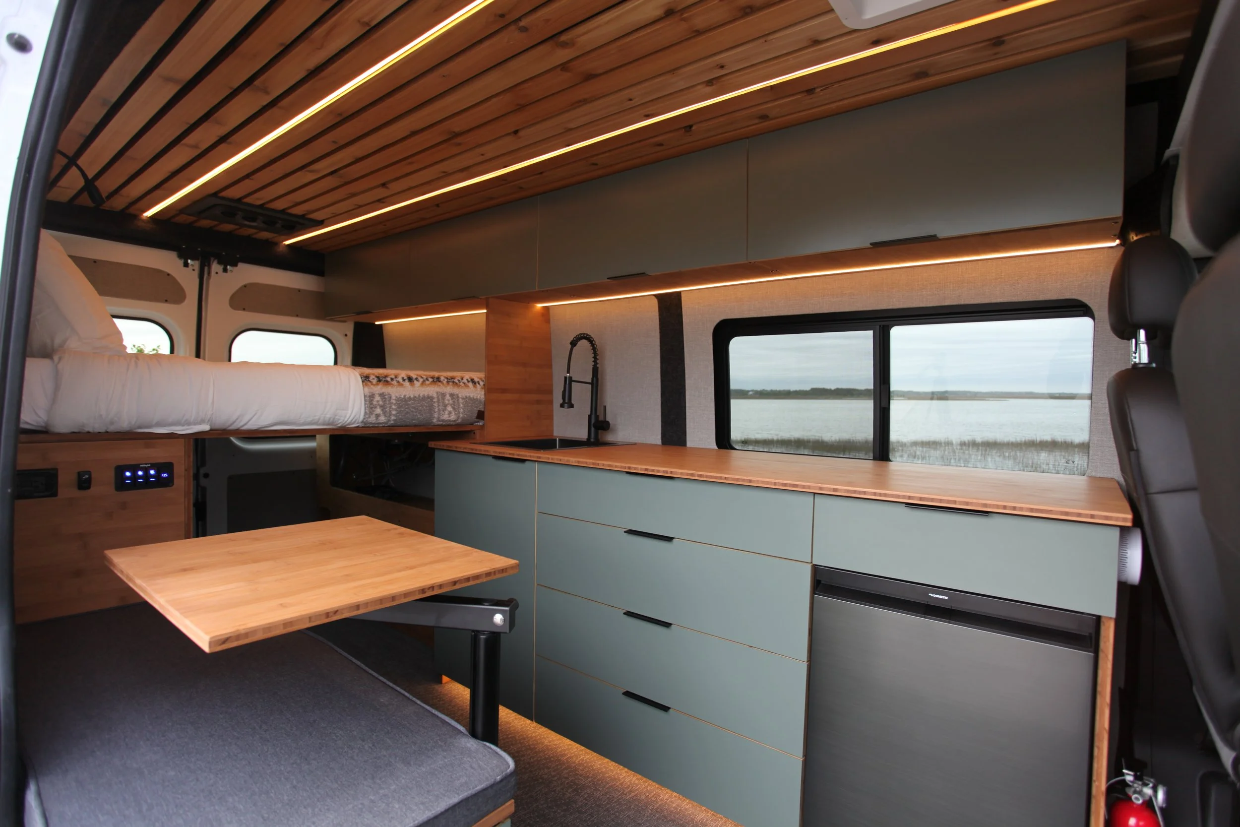 Interior of a converted van with a wooden ceiling, blue cabinets, a countertop with a sink, a window showing water and land outside, and a bed area with white linens.