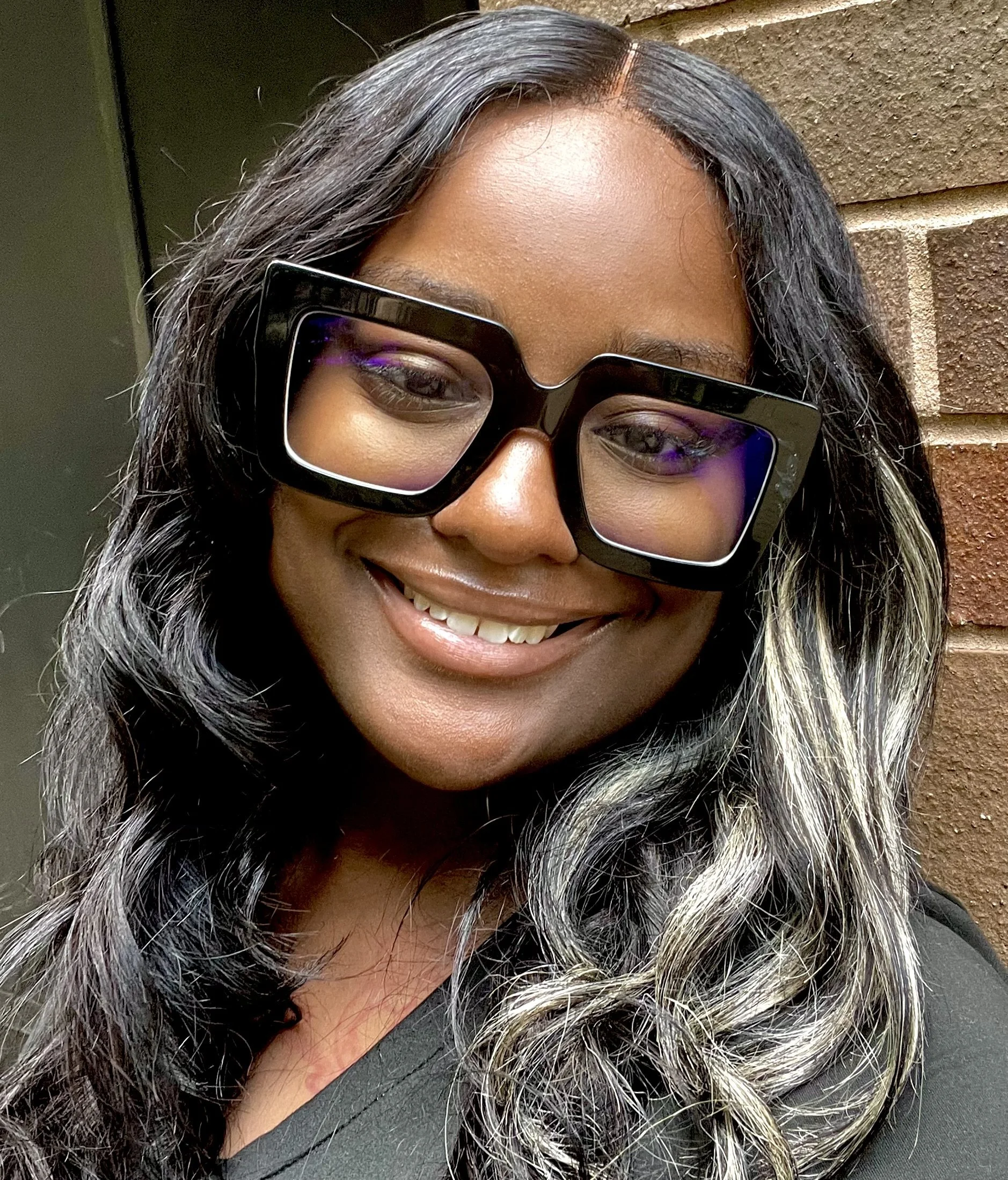 Close-up of a woman with long, wavy hair wearing large black glasses, smiling in front of a brick wall.
