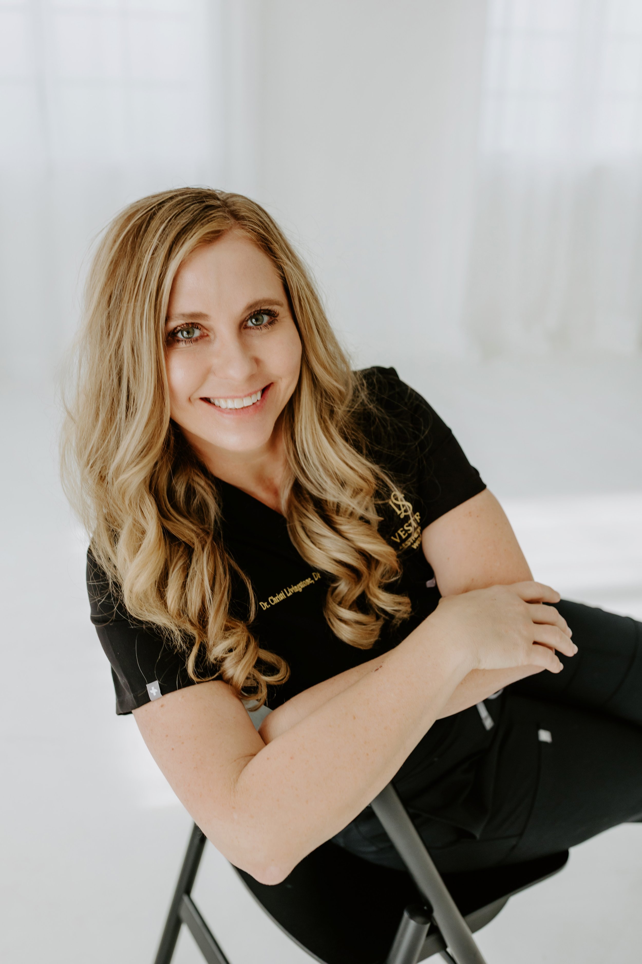 A woman with long blond curly hair smiling while sitting in a black chair, wearing a black uniform with gold embroidery.