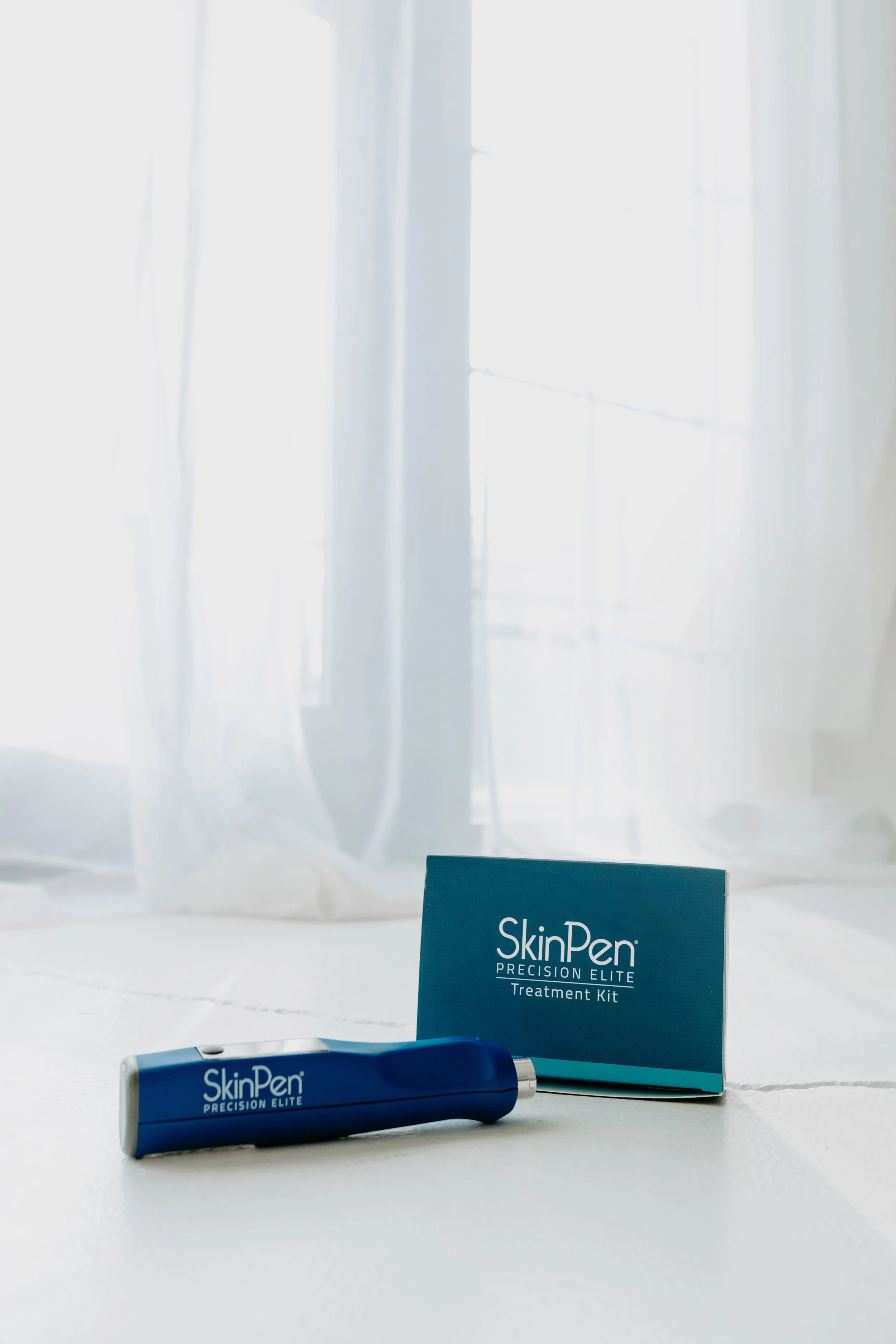 SkinPen Precision Elite treatment kit and device on a white surface near a window with sheer curtains.