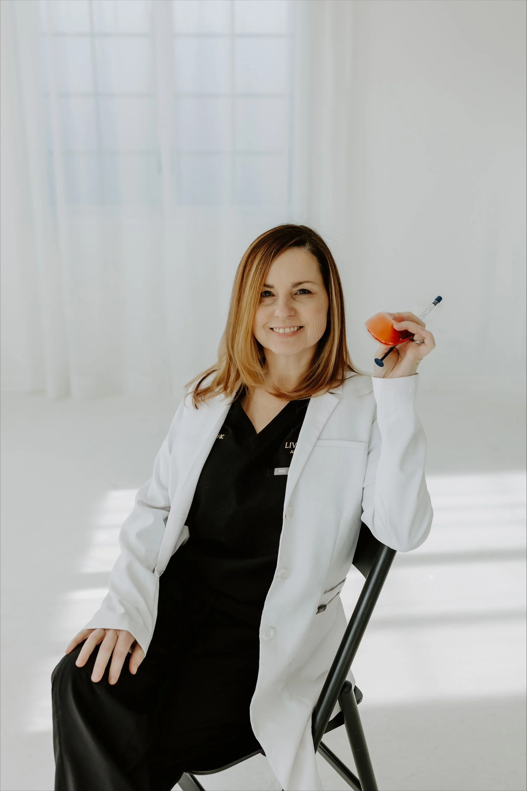 A woman with shoulder-length brown hair wearing a white medical coat sitting on a black chair, holding a syringe with red liquid, smiling in a bright room with large windows.