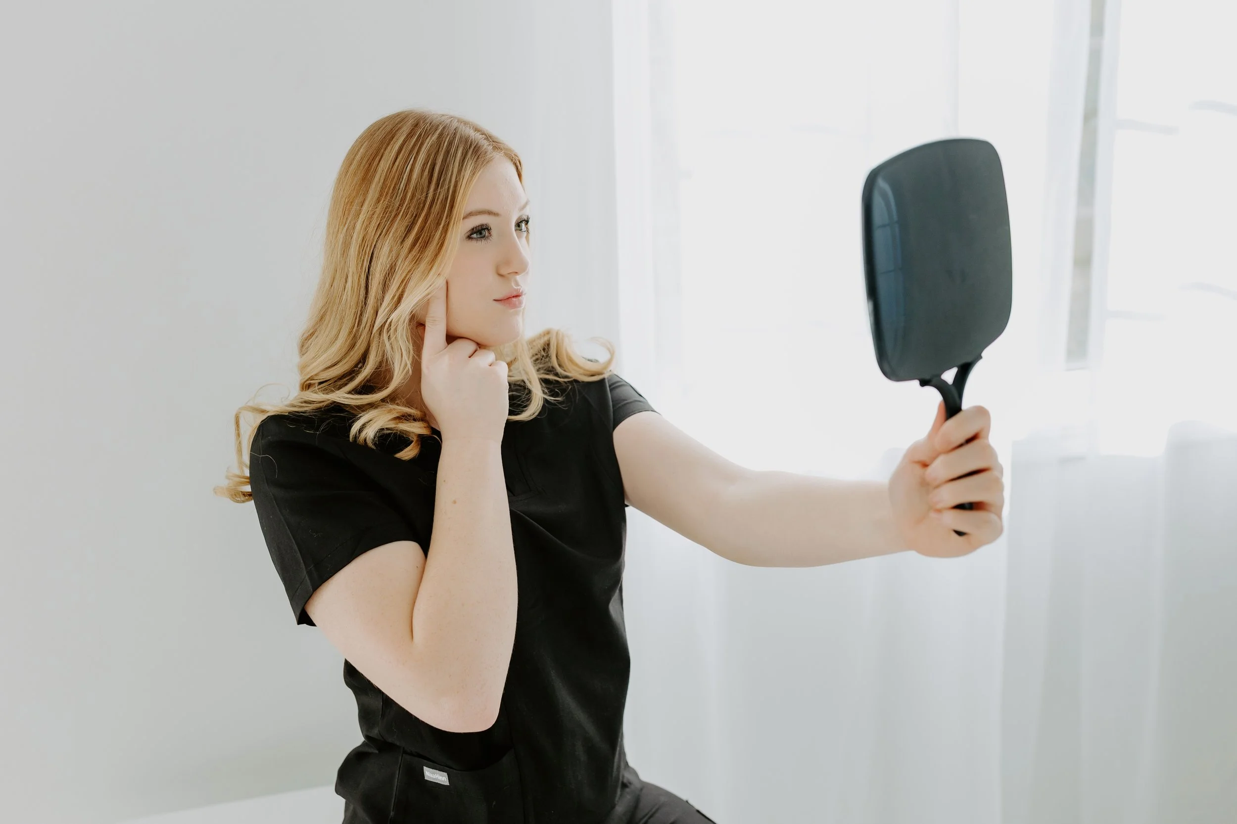 A woman with long blond hair wearing a black shirt and black pants, looking at herself in a handheld mirror with a thoughtful expression, standing in a bright room with white walls and sheer curtains.