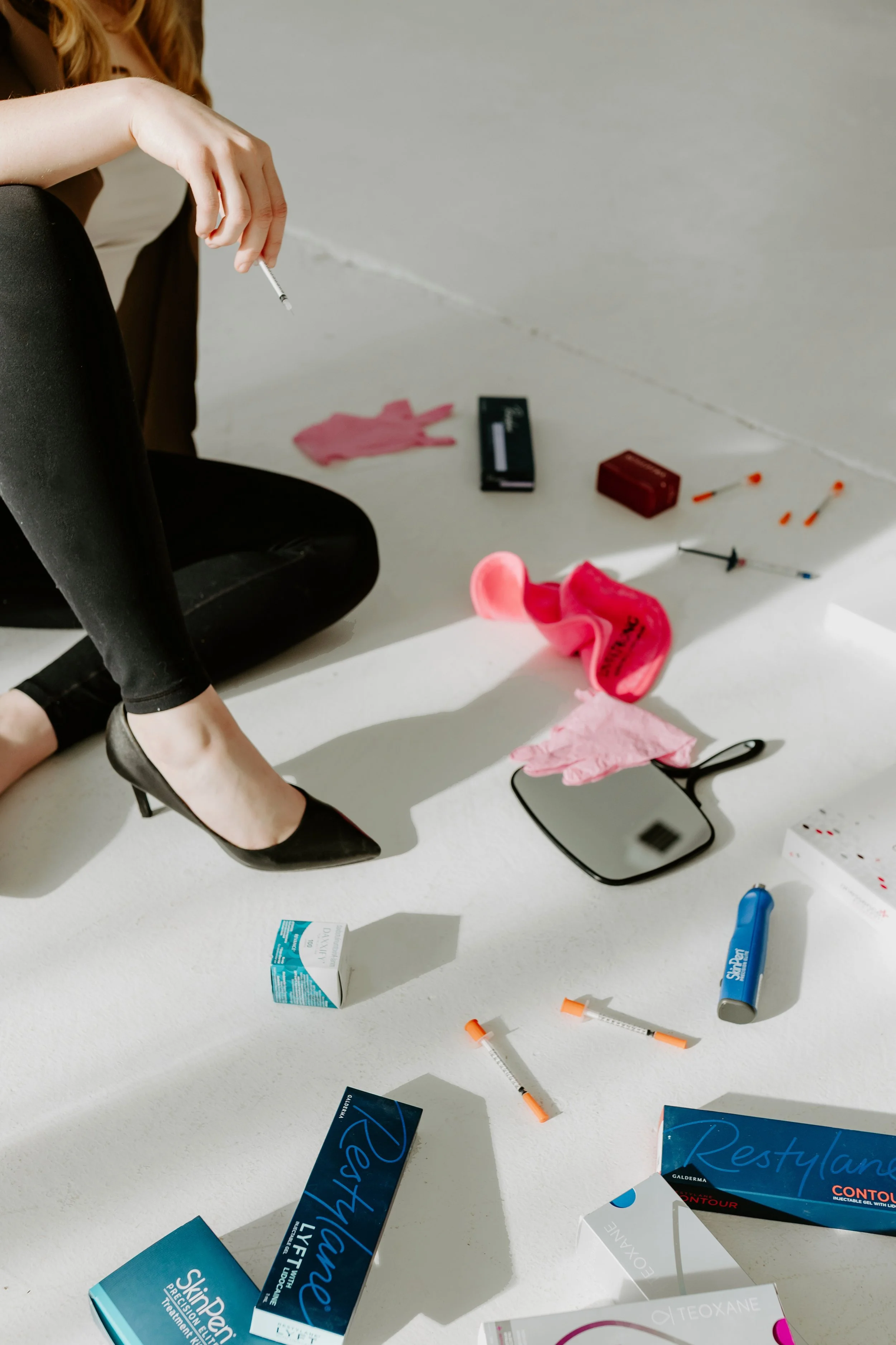 Disarrayed pills, syringes, and medication boxes scattered on a white floor with a person sitting nearby.