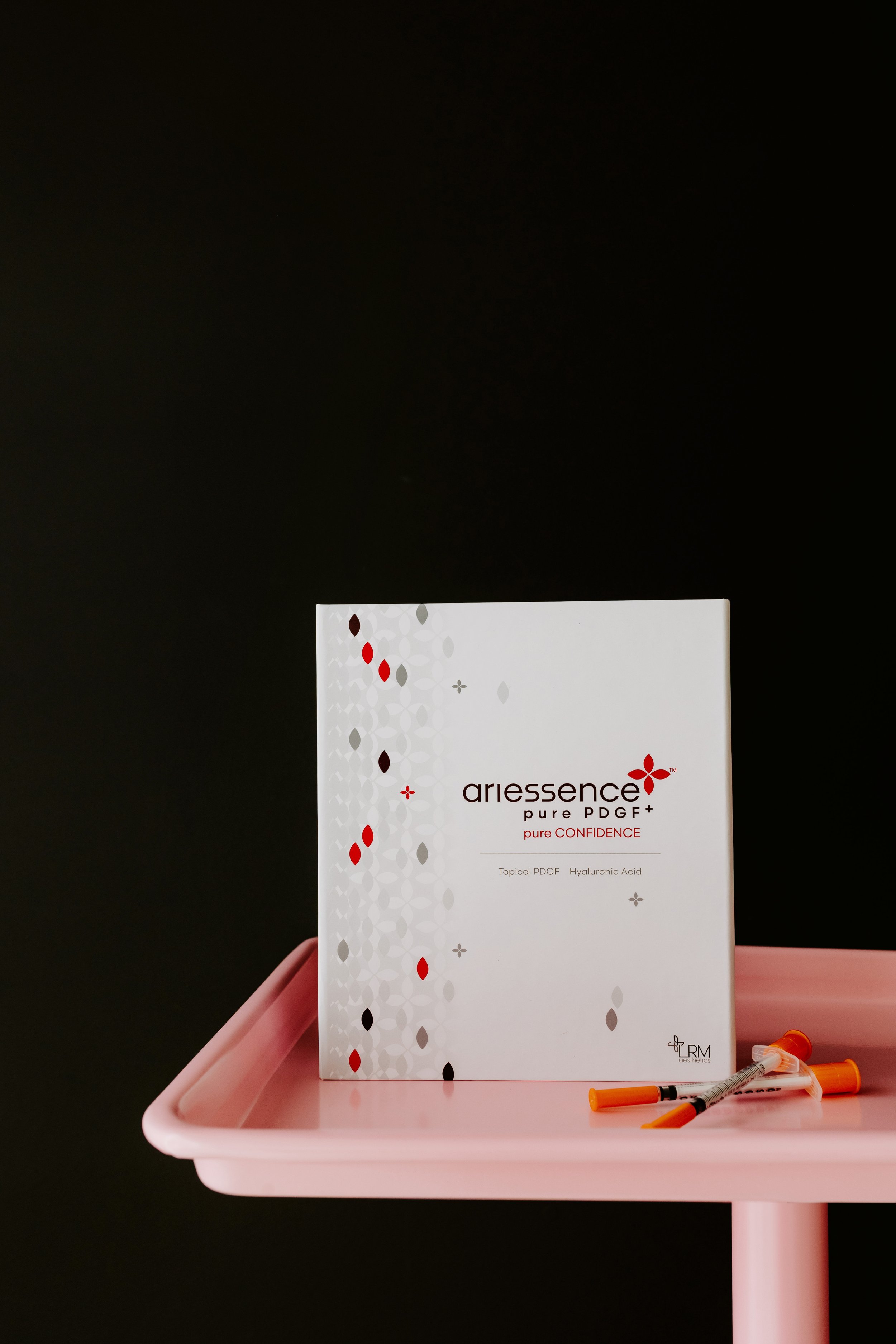 A box of Ariessence Pure PDGF+ skincare product with syringes and vials on a pink tray against a black background.