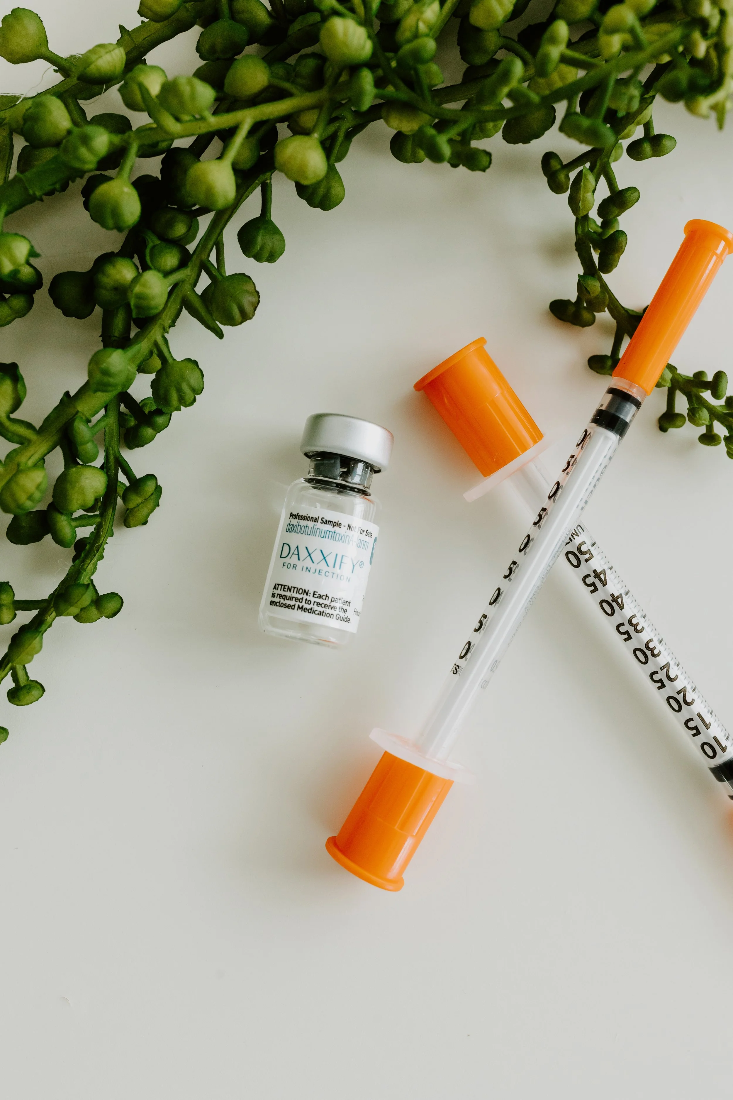 A small vial labeled DAXXIFY with a medical syringe, and an orange cap, on a white surface with green sprigs of plants nearby.