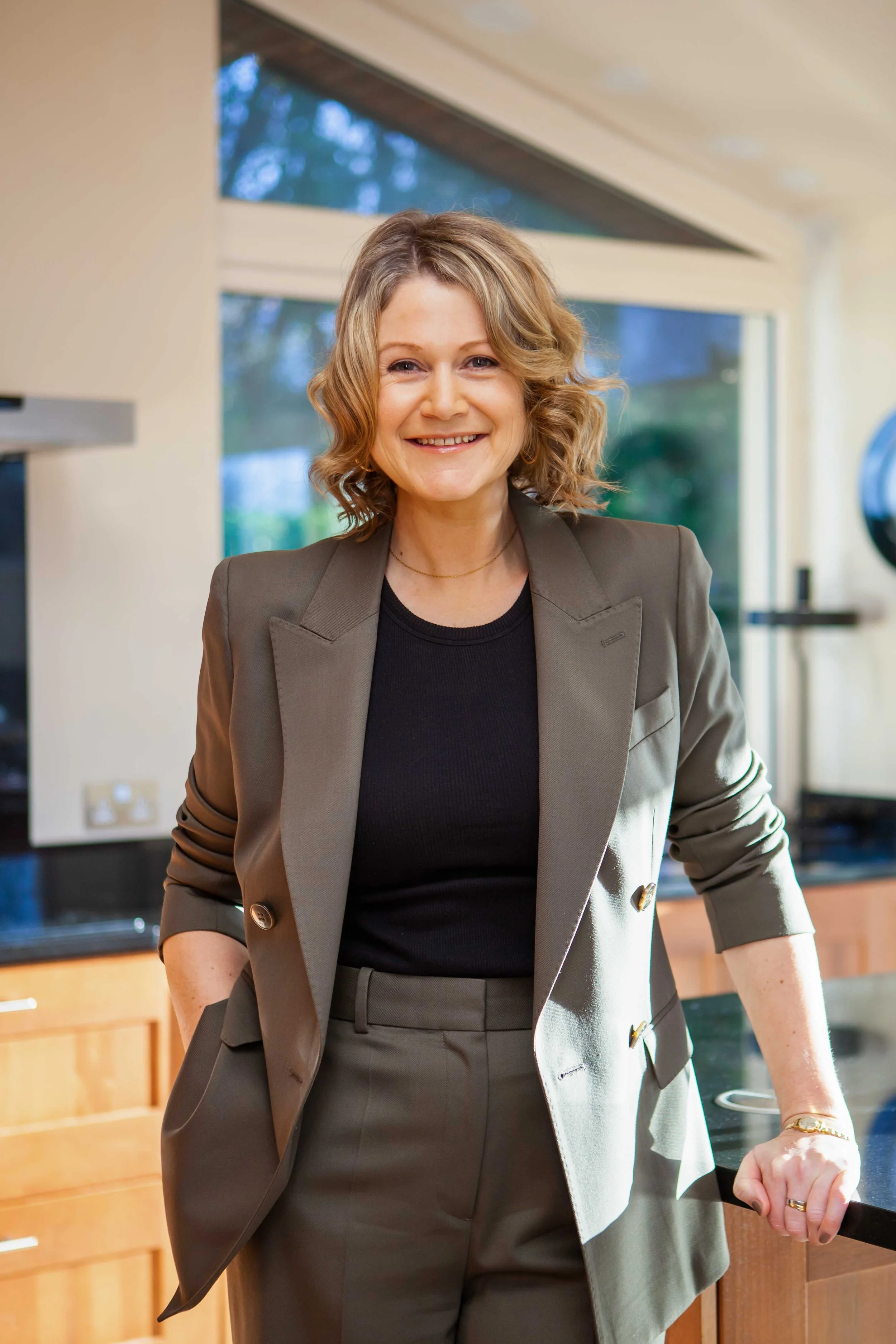 A woman with curly blonde hair smiling in a modern kitchen, wearing a khaki suit and black top.