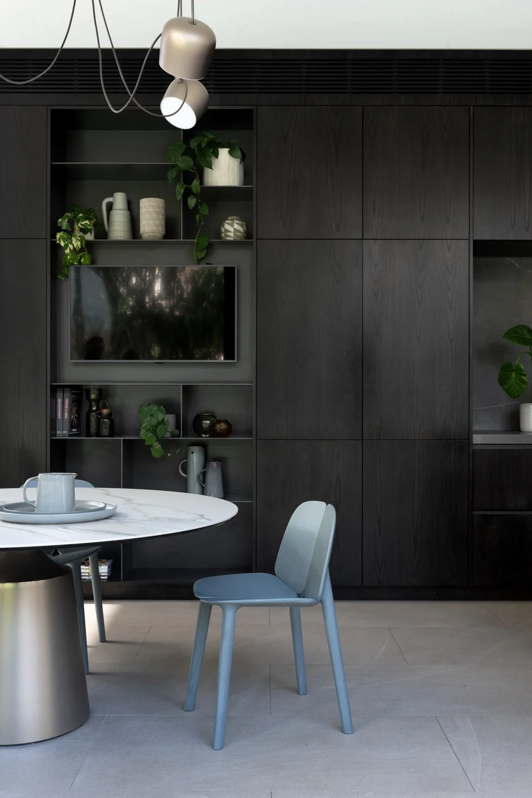 Modern living room with a black built-in cabinet, a wall-mounted TV, and decorative vases and plants, with a round white marble table and a light blue chair in the foreground.