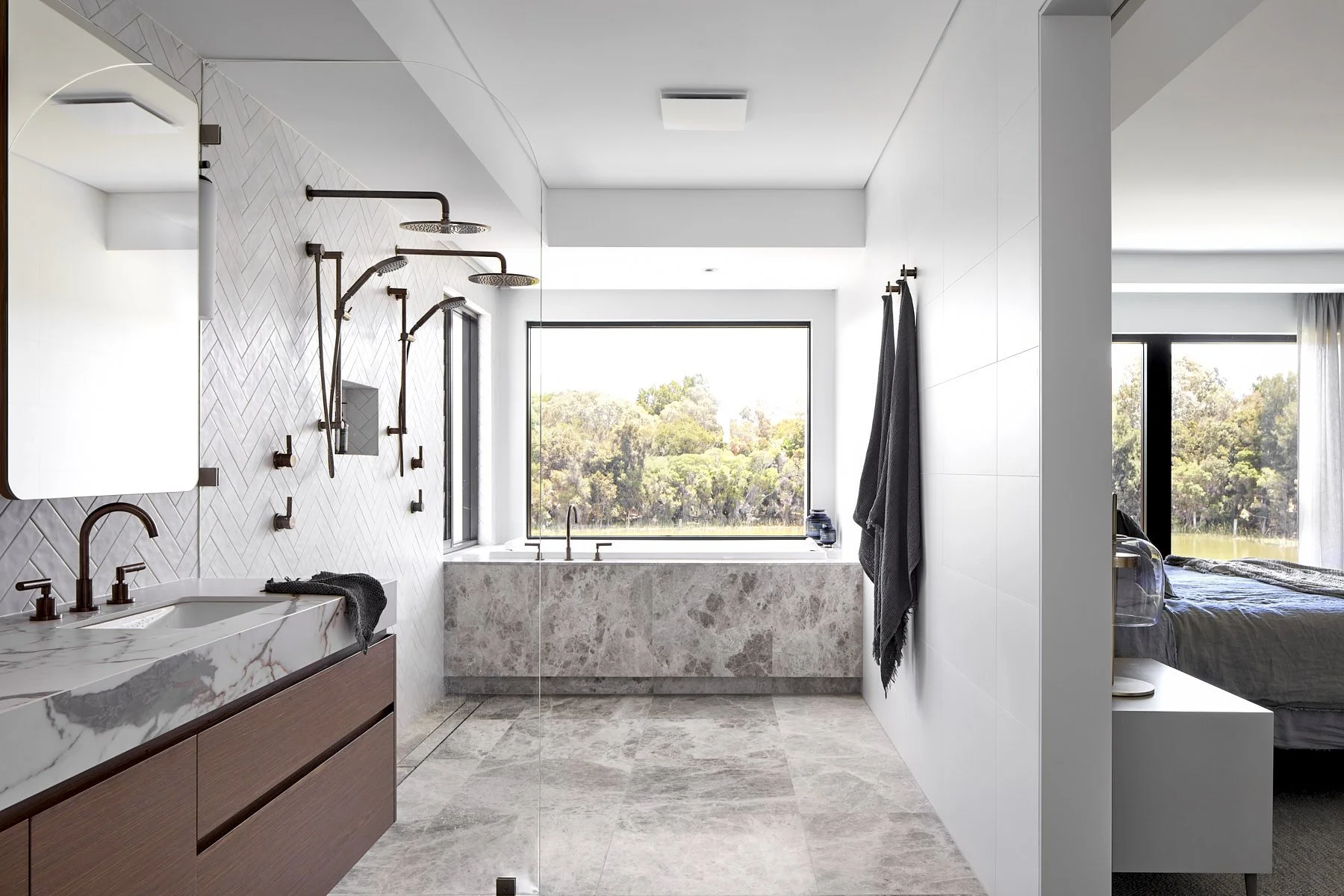 Modern bathroom with a marble vanity, two shower heads, a large window, and a separate bedroom visible through an open wall.