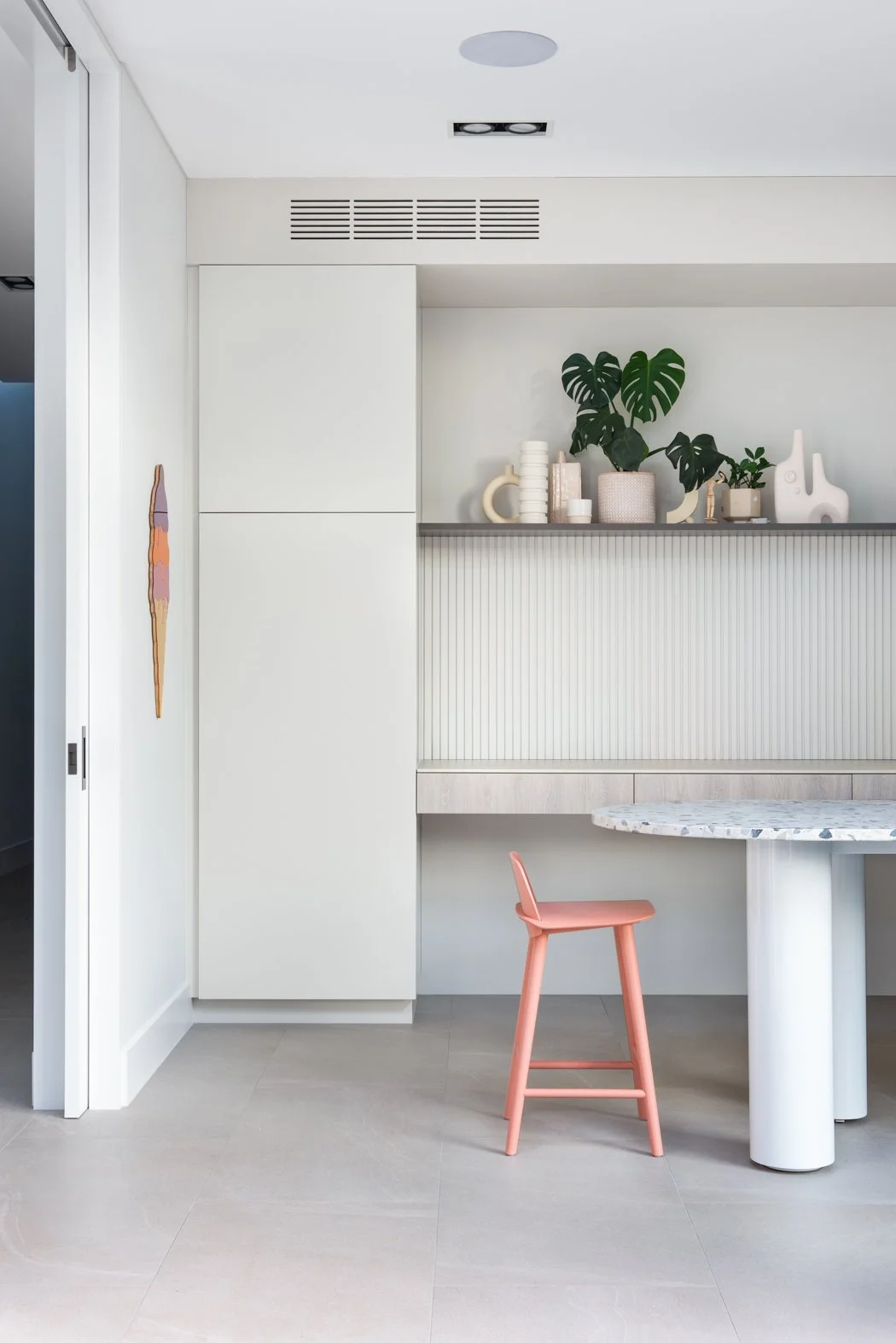 Modern, minimalist art studio with white cabinetry, a marble tabletop, pink barstool, and decorative pottery and plants on a shelf.
