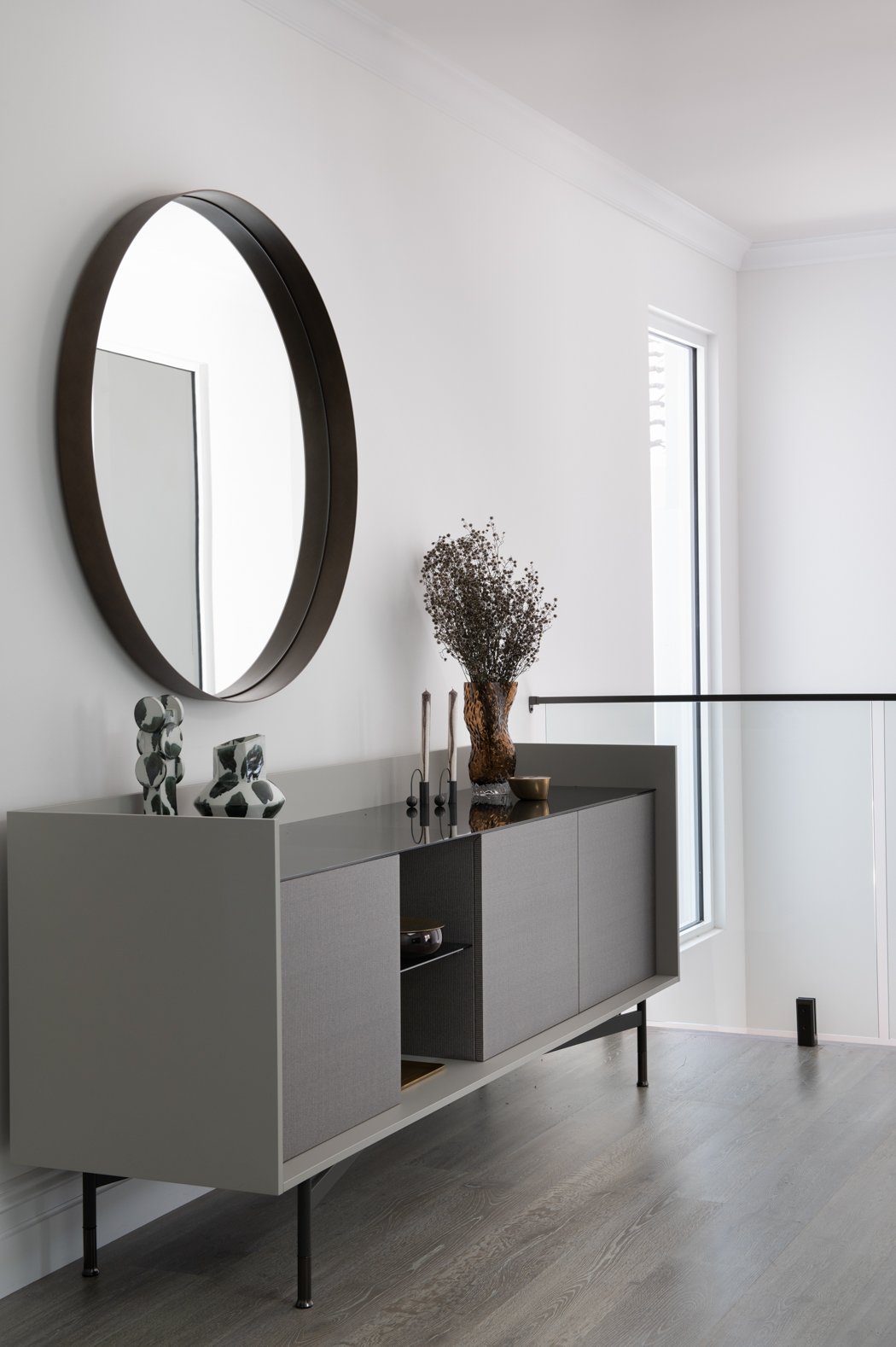 Modern interior with a black framed oval mirror above a grey sideboard decorated with black and white vases, a brown vase with dried flowers, and small decorative objects, next to a large window with natural light.