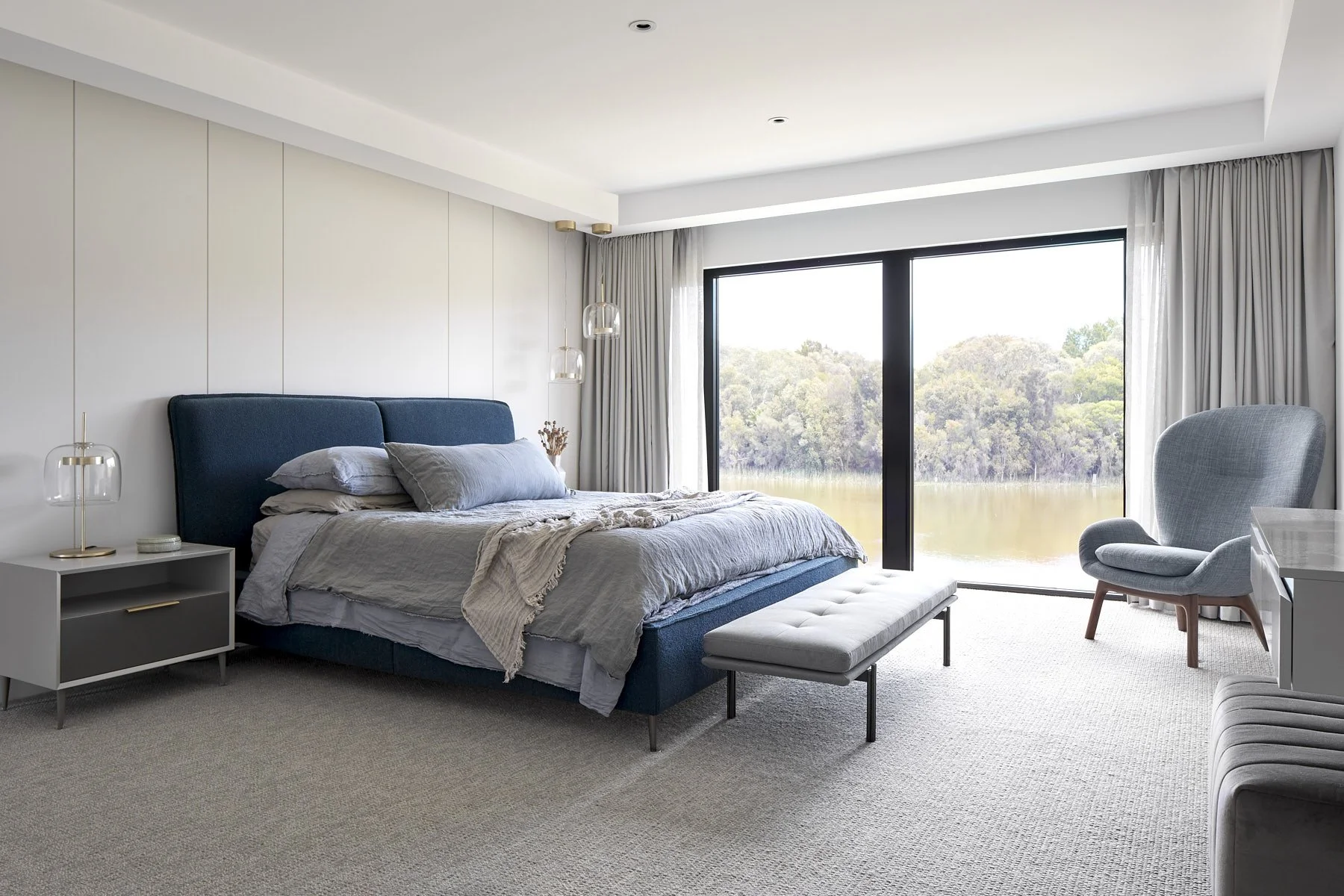 Modern bedroom with large window overlooking a lake and trees, blue upholstered bed, blue bedding, light blue bench at foot of bed, blue armchair near window, side table with lamps.