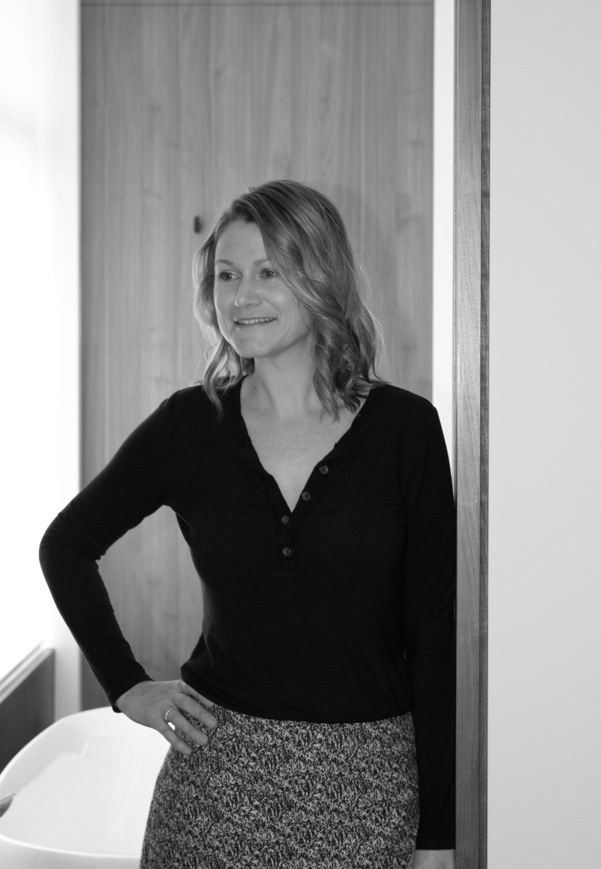 A woman with shoulder-length hair, wearing a black button-up top and patterned skirt, standing indoors near a wooden wall and smiling.