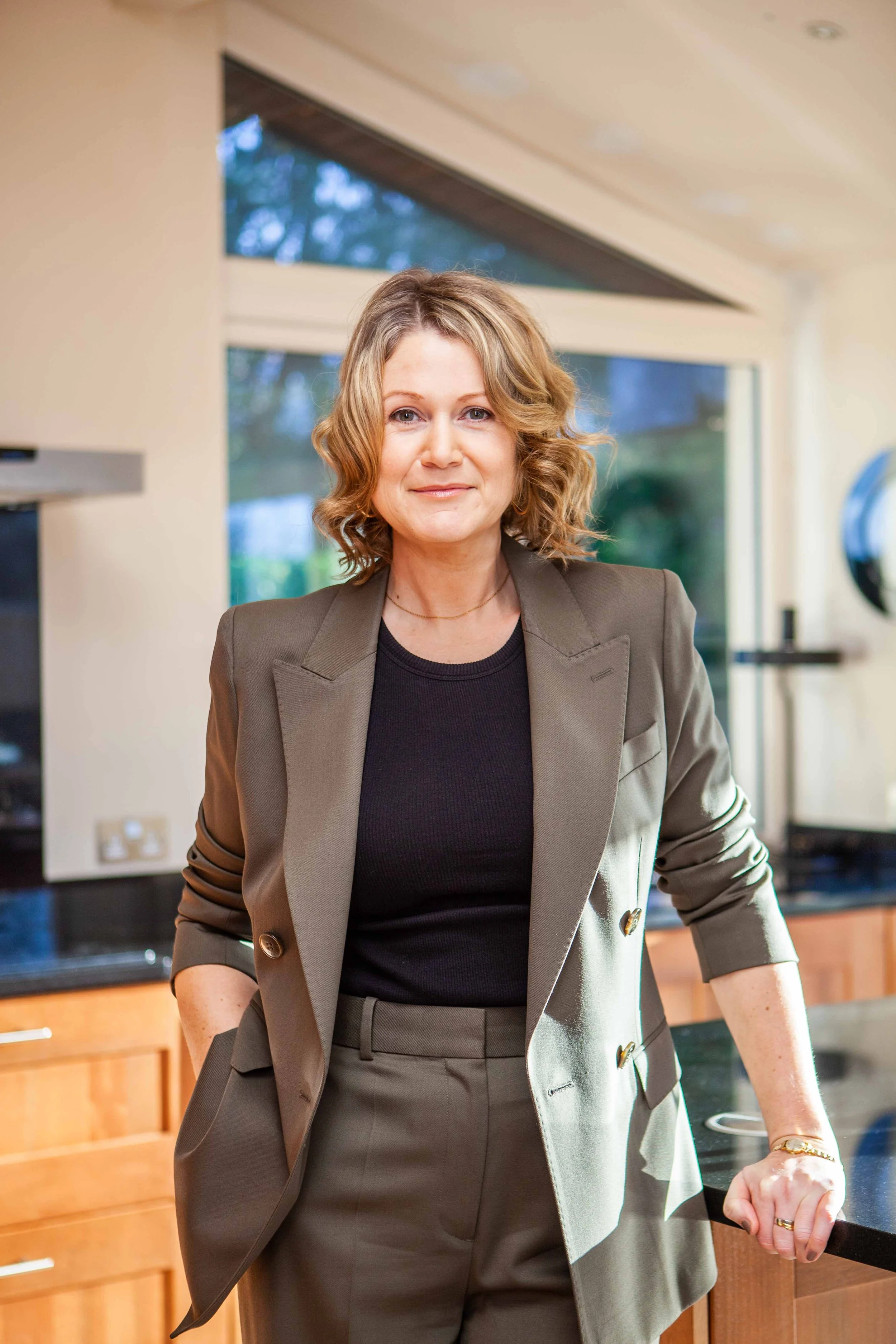A woman with curly blonde hair, wearing a khaki blazer and black top, standing in a kitchen with wooden cabinets and large windows.