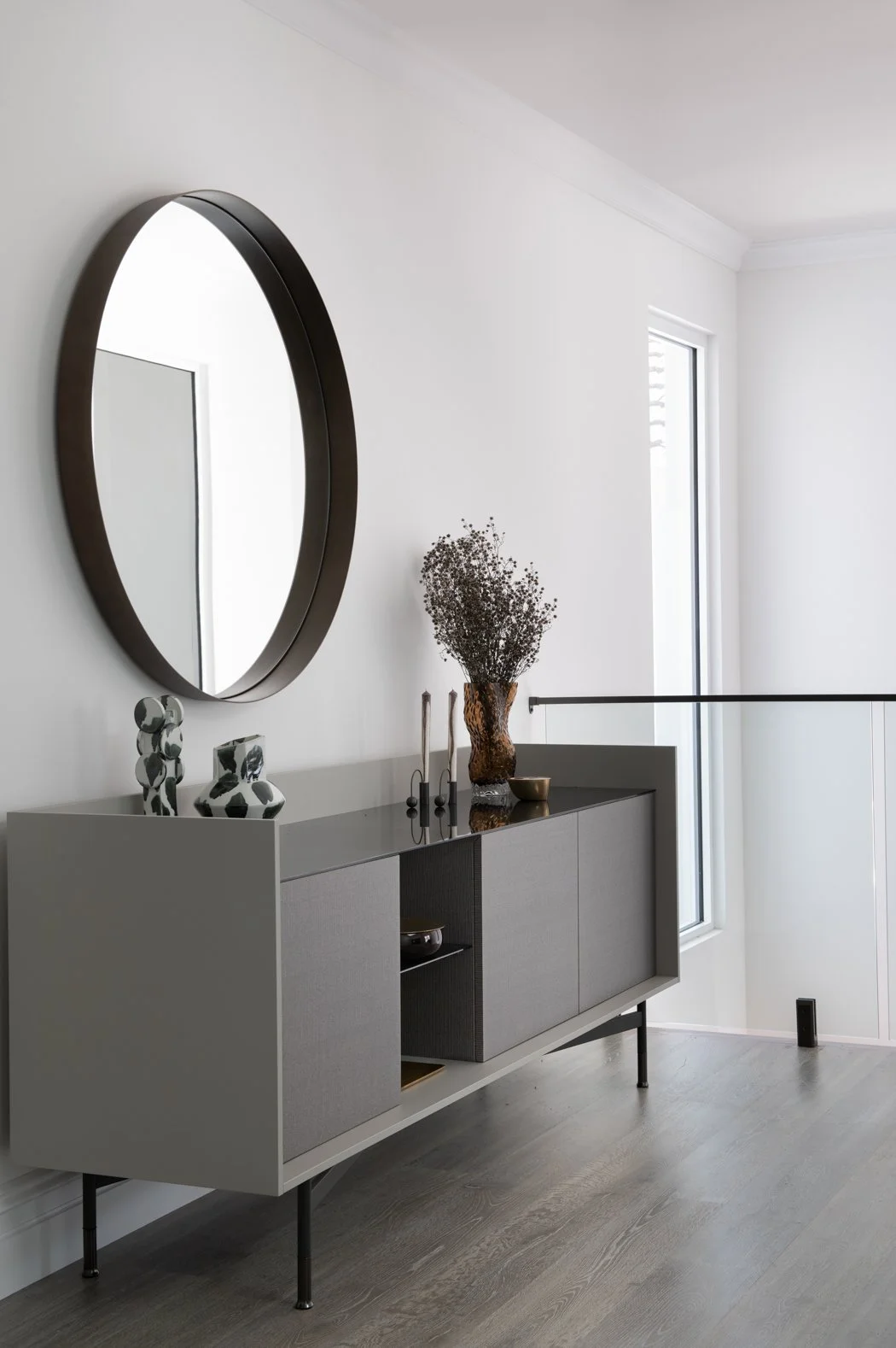 Modern grey sideboard with decorative vases, candles, and a dried flower arrangement, mounted oval mirror, in a bright corner with a large window.