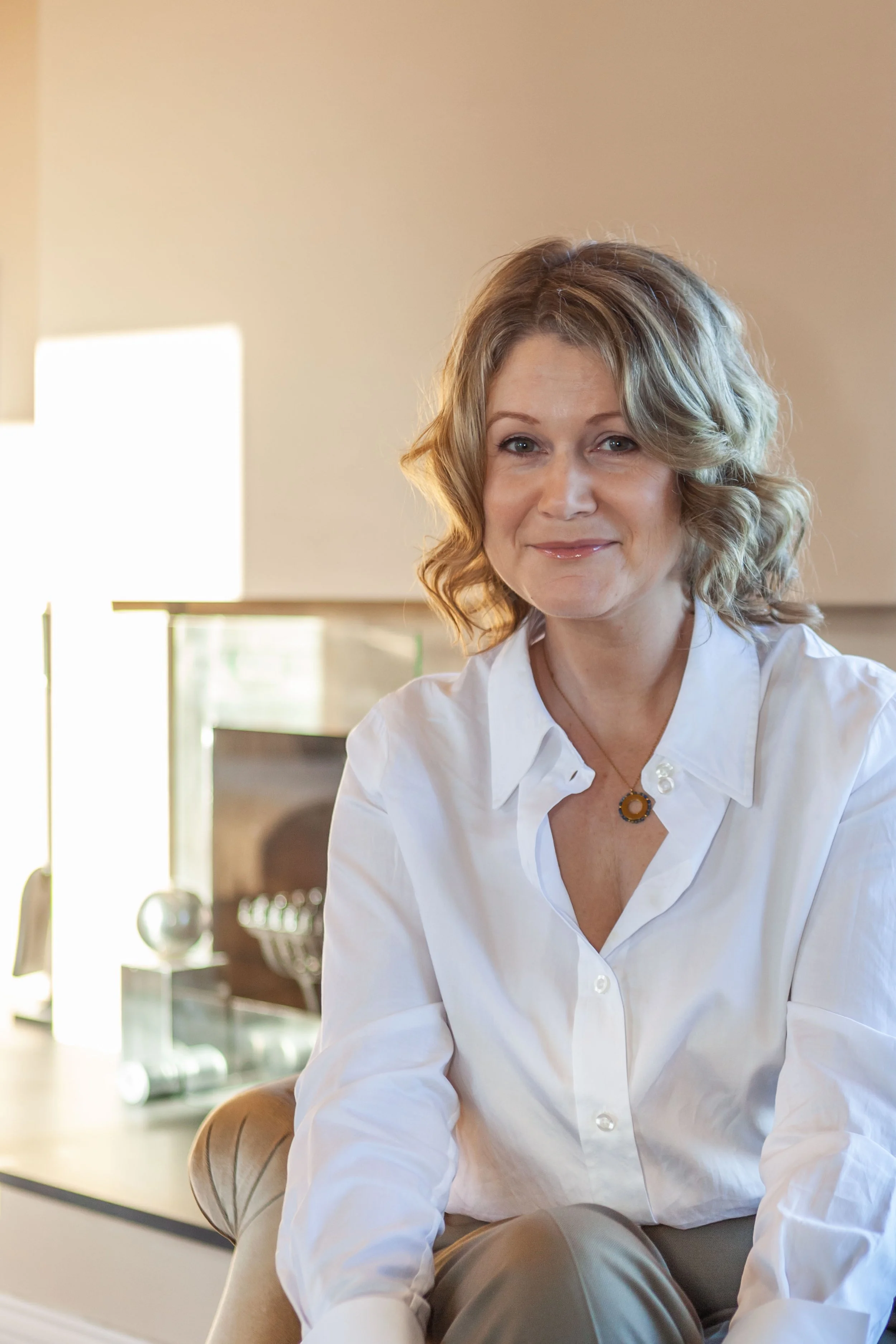 A woman with shoulder-length blonde curly hair, wearing a white button-up shirt and a necklace, sitting on a chair. The background includes a kitchen setting with a sink, a glass container with utensils, and some sunlight coming in.