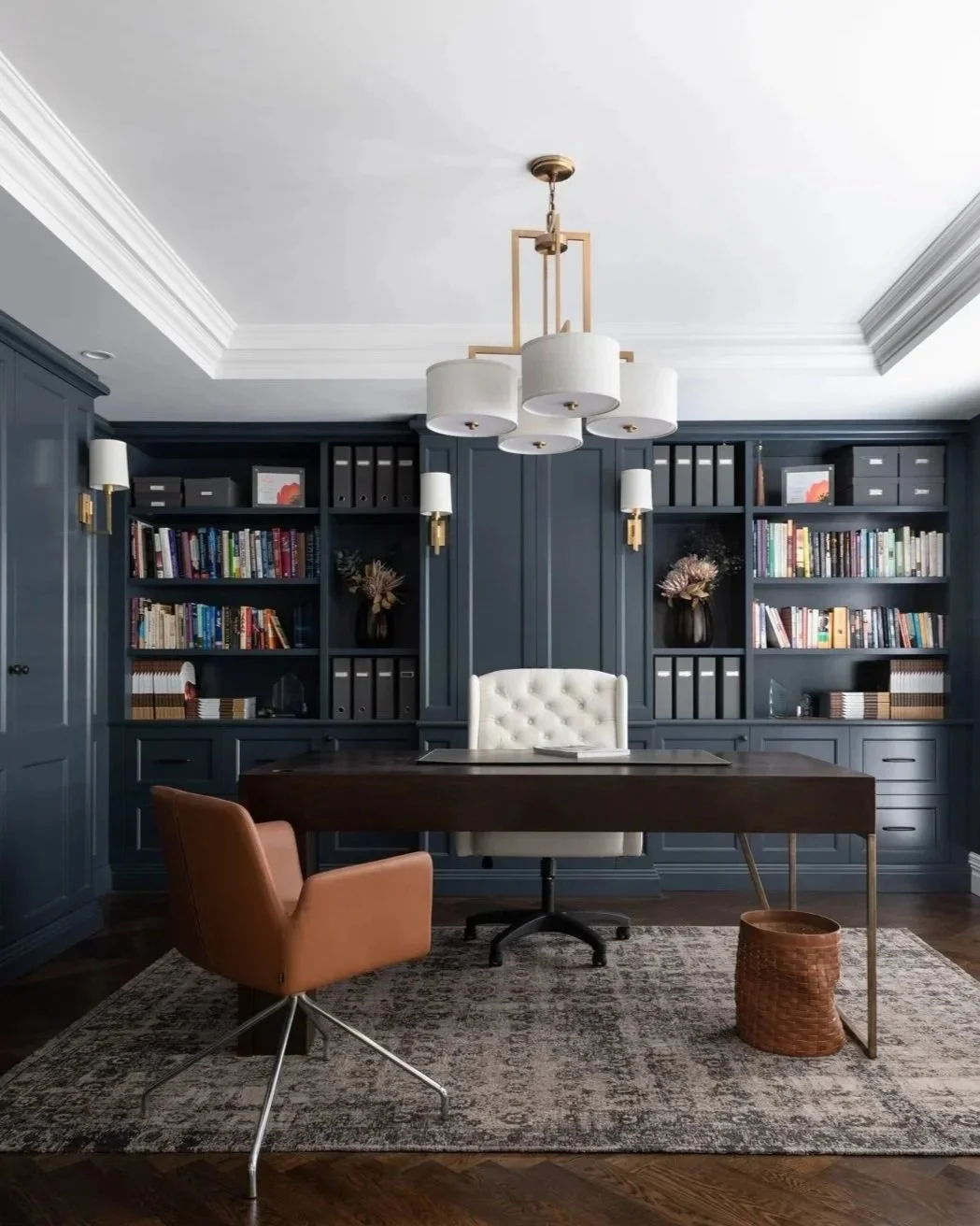 Home office with navy blue built-in bookshelf behind a large dark wood desk, a white tufted office chair, and a tan accent chair on a patterned rug under a modern chandelier.