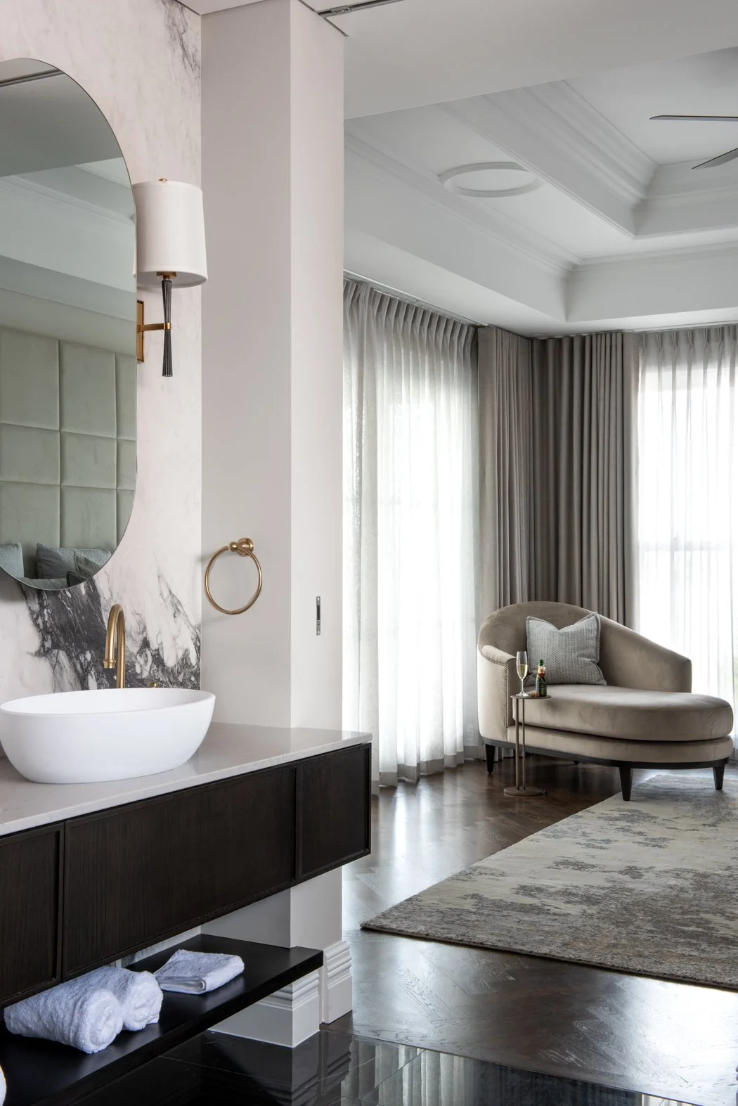 A luxurious bedroom features a curved chaise lounge with a pillow and a side table, large floor-to-ceiling windows with sheer curtains, and a sleek bathroom vanity with a vessel sink, gold fixtures, and a large mirror on a marble wall.