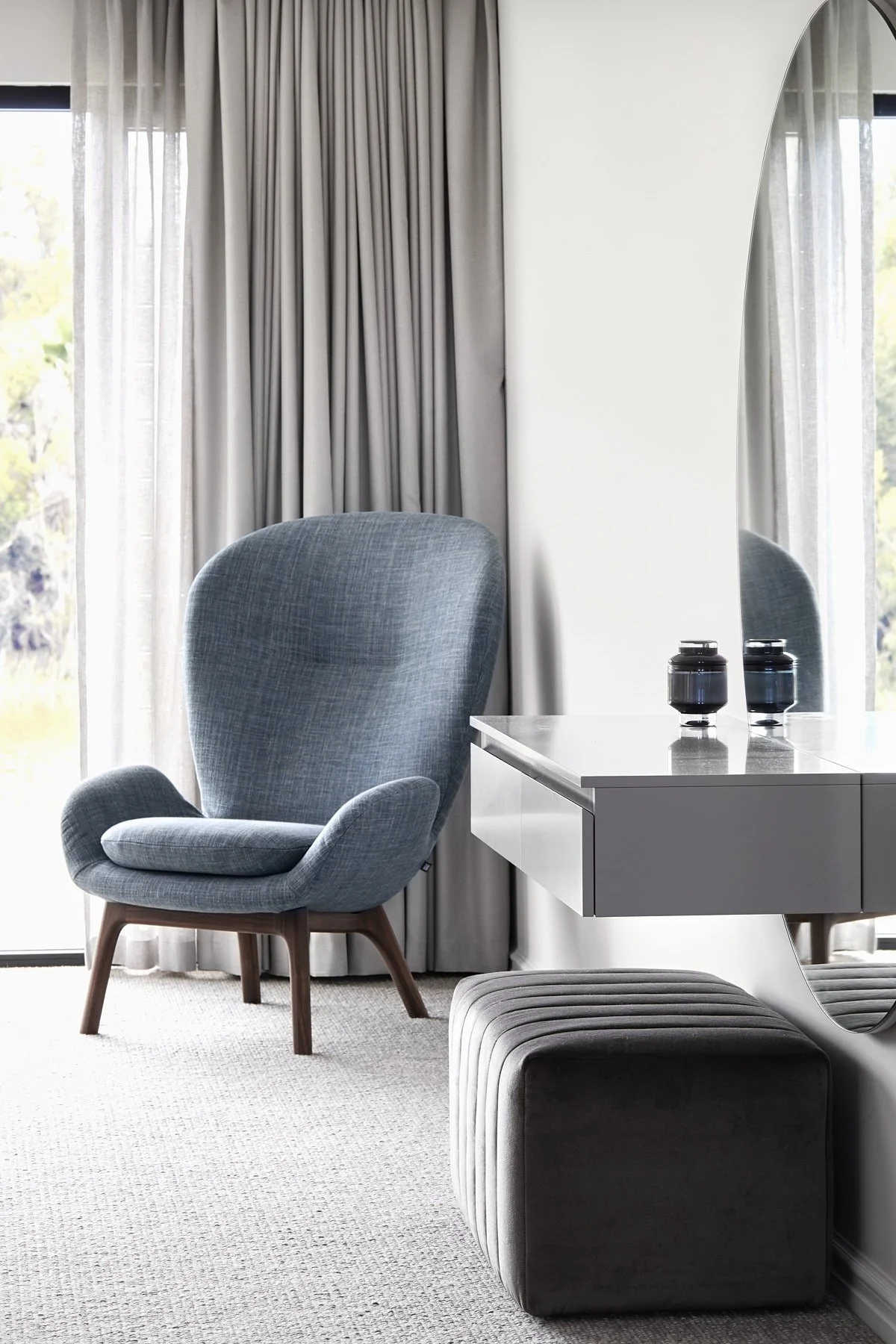 A modern bedroom corner with a blue upholstered armchair, a blue floating vanity with two black vases, a blue ottoman, and floor-to-ceiling windows with light curtains.