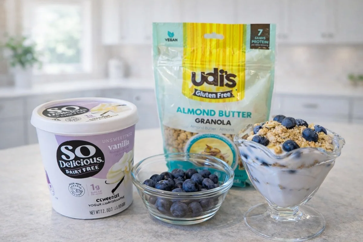Ingredients for a dairy-free yogurt parfait, including dairy-free yogurt, gluten-free granola, blueberries, and a prepared parfait in a glass dish.