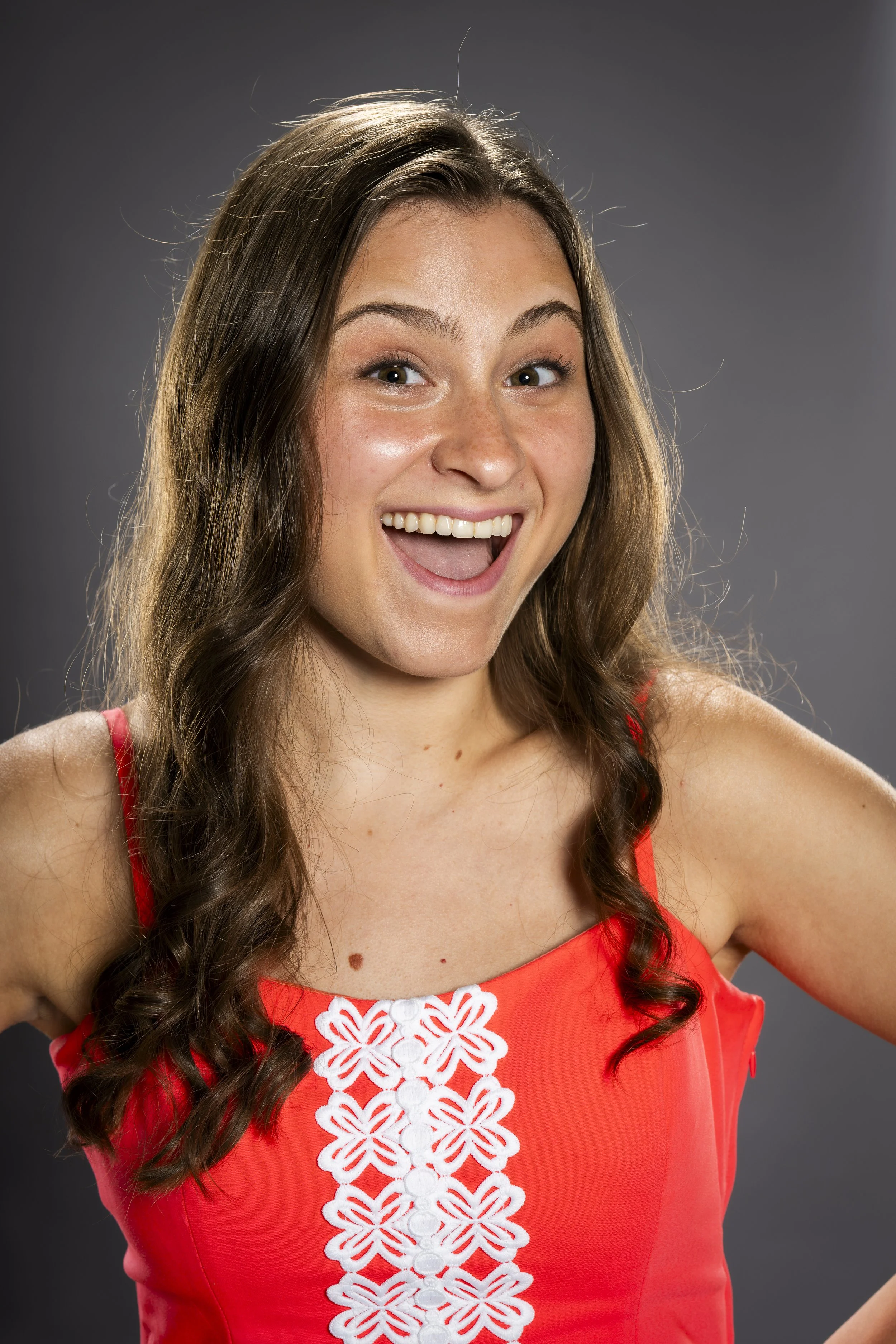 A young woman with long, wavy brown hair smiling and looking excited, wearing a red sleeveless top with white floral embroidery in the center.