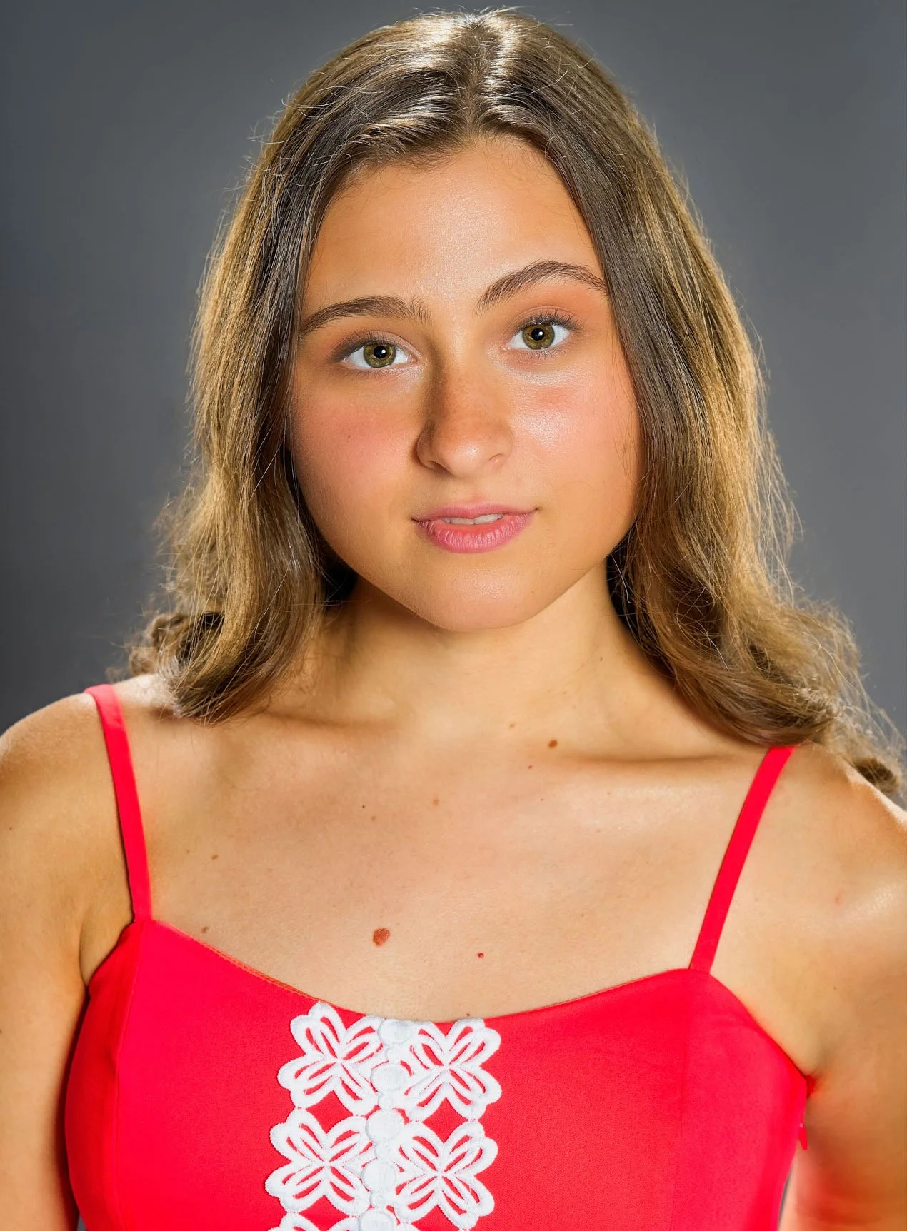 A young woman with long brown hair, light skin, and green eyes, wearing a red spaghetti strap top with white embroidery, looking directly at the camera with a neutral expression.