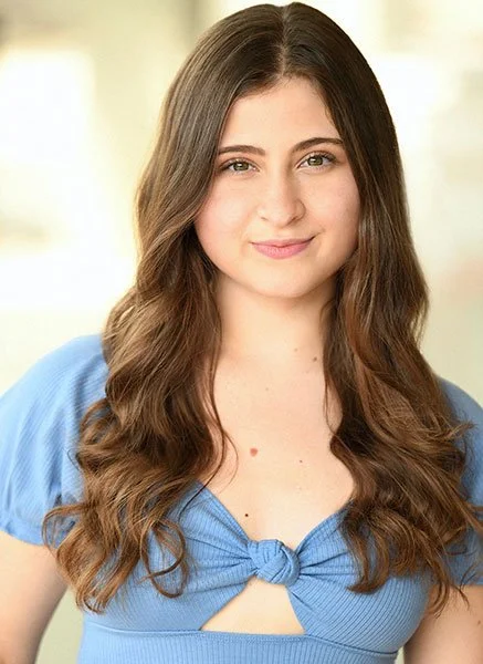 Young woman with long wavy brown hair wearing a blue top with a knot in the front, smiling softly, in a bright indoor setting.