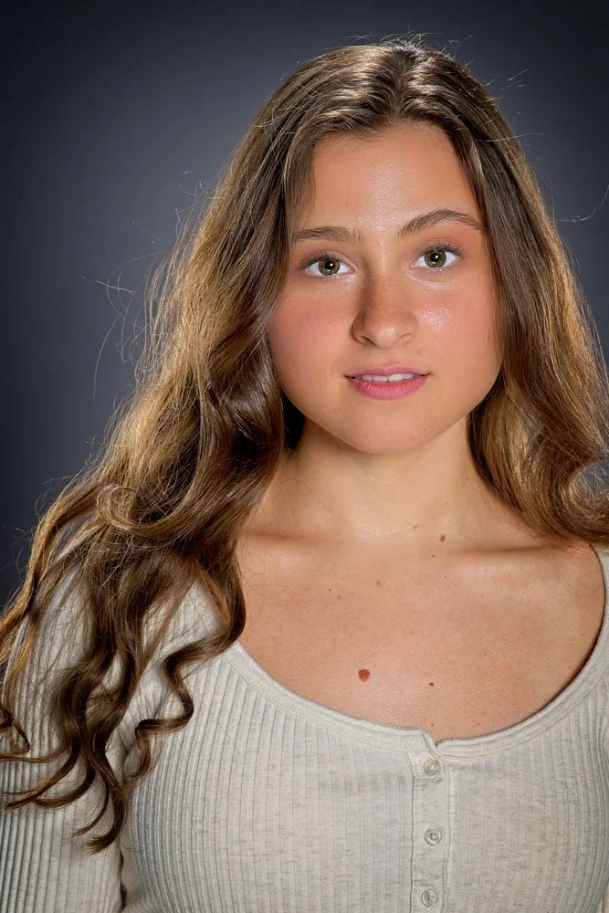 A young woman with long, wavy brown hair and green eyes, wearing a white ribbed long-sleeve shirt, looking at the camera, against a dark gray background.