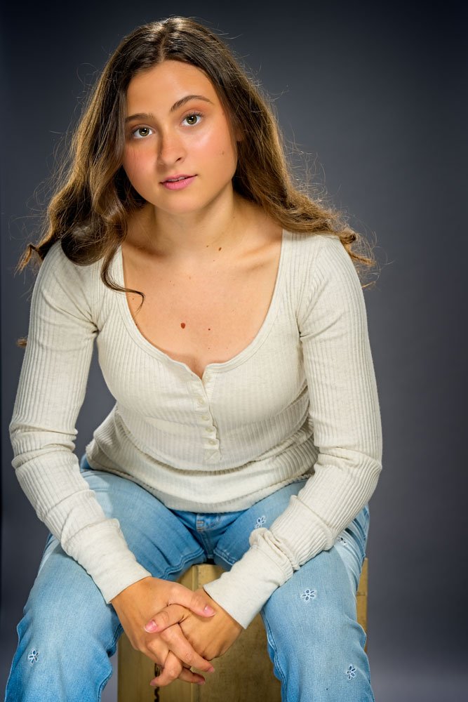 Young woman with long, wavy brown hair sitting on a wooden stool against a dark background, wearing a cream ribbed long-sleeve shirt and blue jeans with embroidered flowers, looking at the camera with a neutral expression.